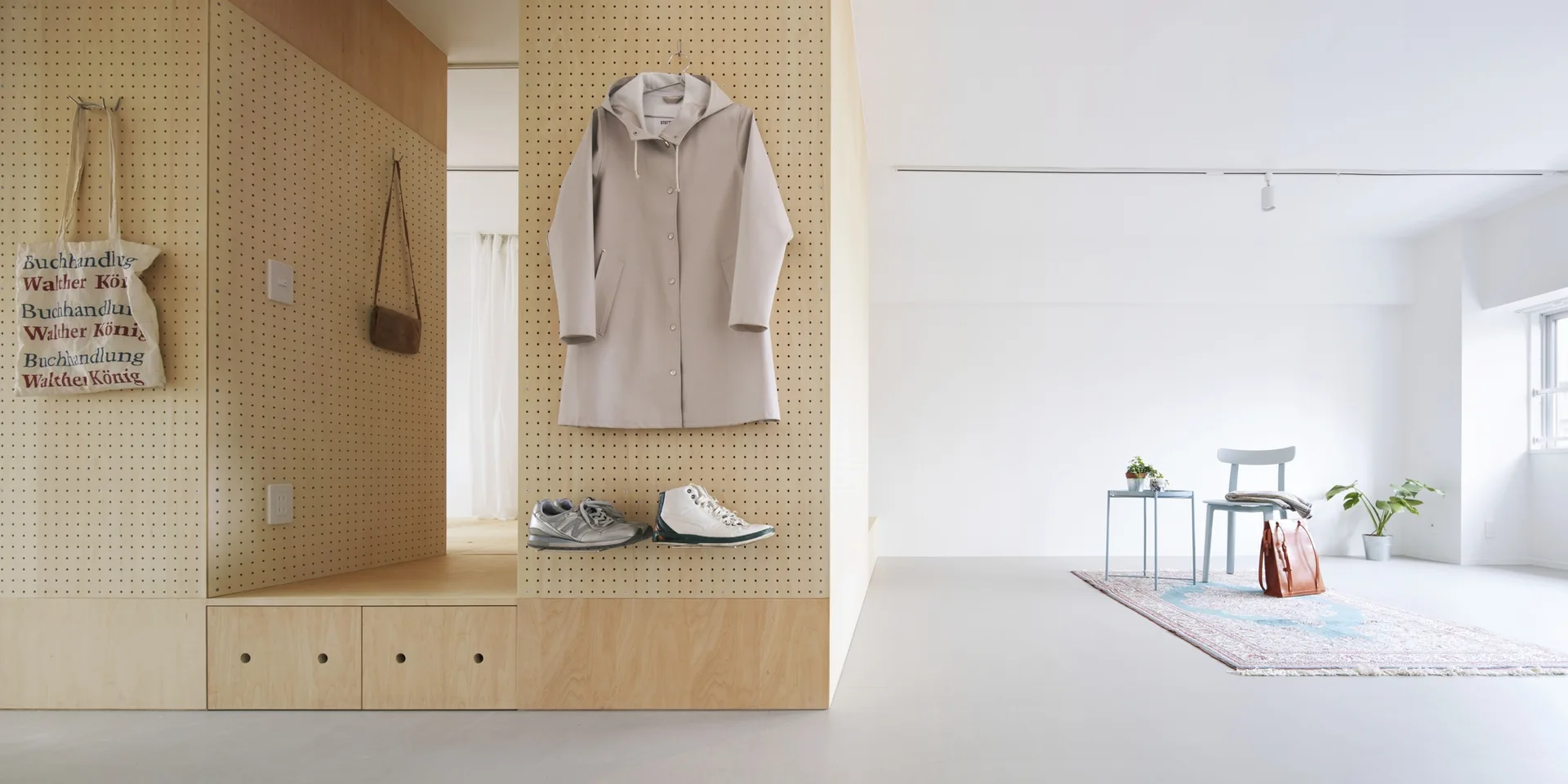 A minimalist room features a plywood pegboard wall divider with hung clothing and shoes, next to a seating area with a blue chair and patterned rug.