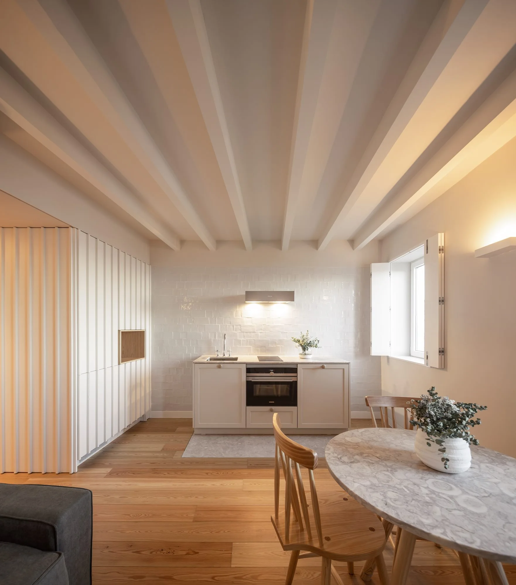 A minimalist kitchen features a compact beige cabinet island, white tiled backsplash, exposed ceiling beams, and a round marble dining table on wood floors.