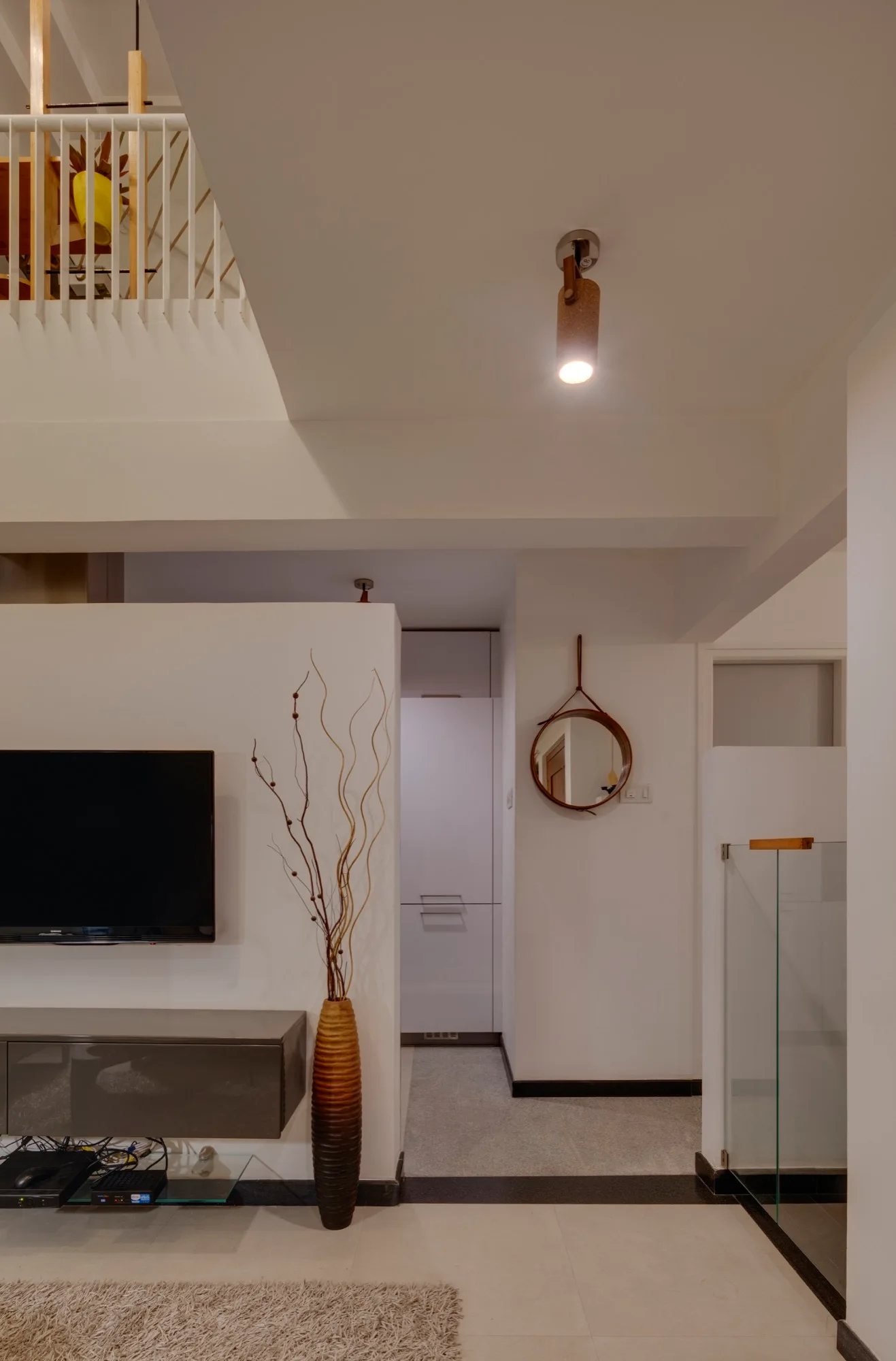 A modern living space features a wall-mounted TV over a floating grey console, a tall decorative floor vase, and a round wall mirror near a staircase.