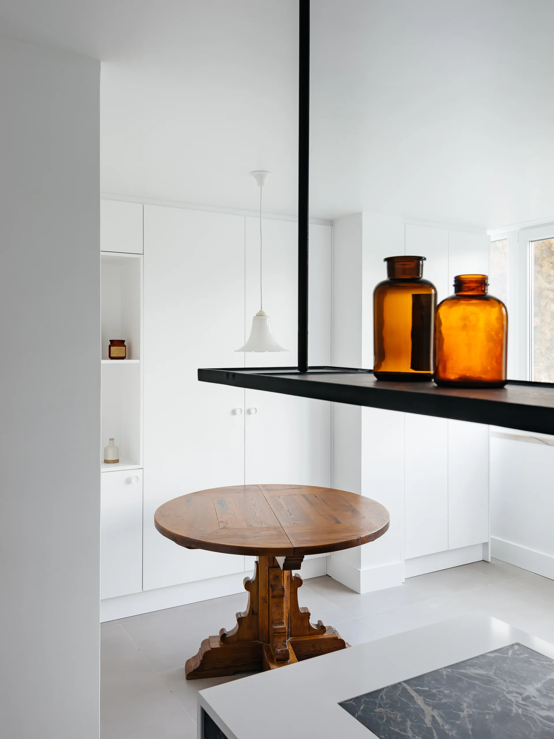 A minimalist white kitchen featuring a round wooden pedestal table, seamless flat-panel cabinetry, and a black suspended shelf with amber glass bottles.