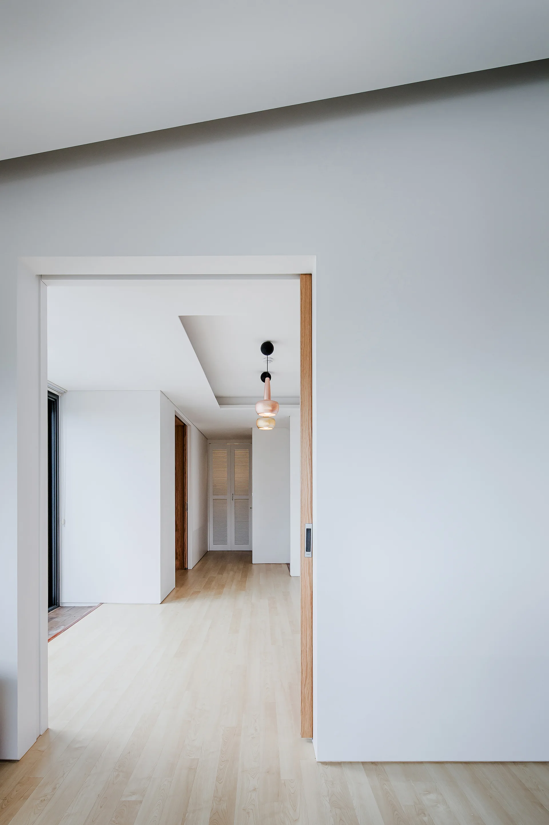An open doorway with a light wood frame looks into a bright minimalist hallway with wood plank flooring and a rose gold pendant light.