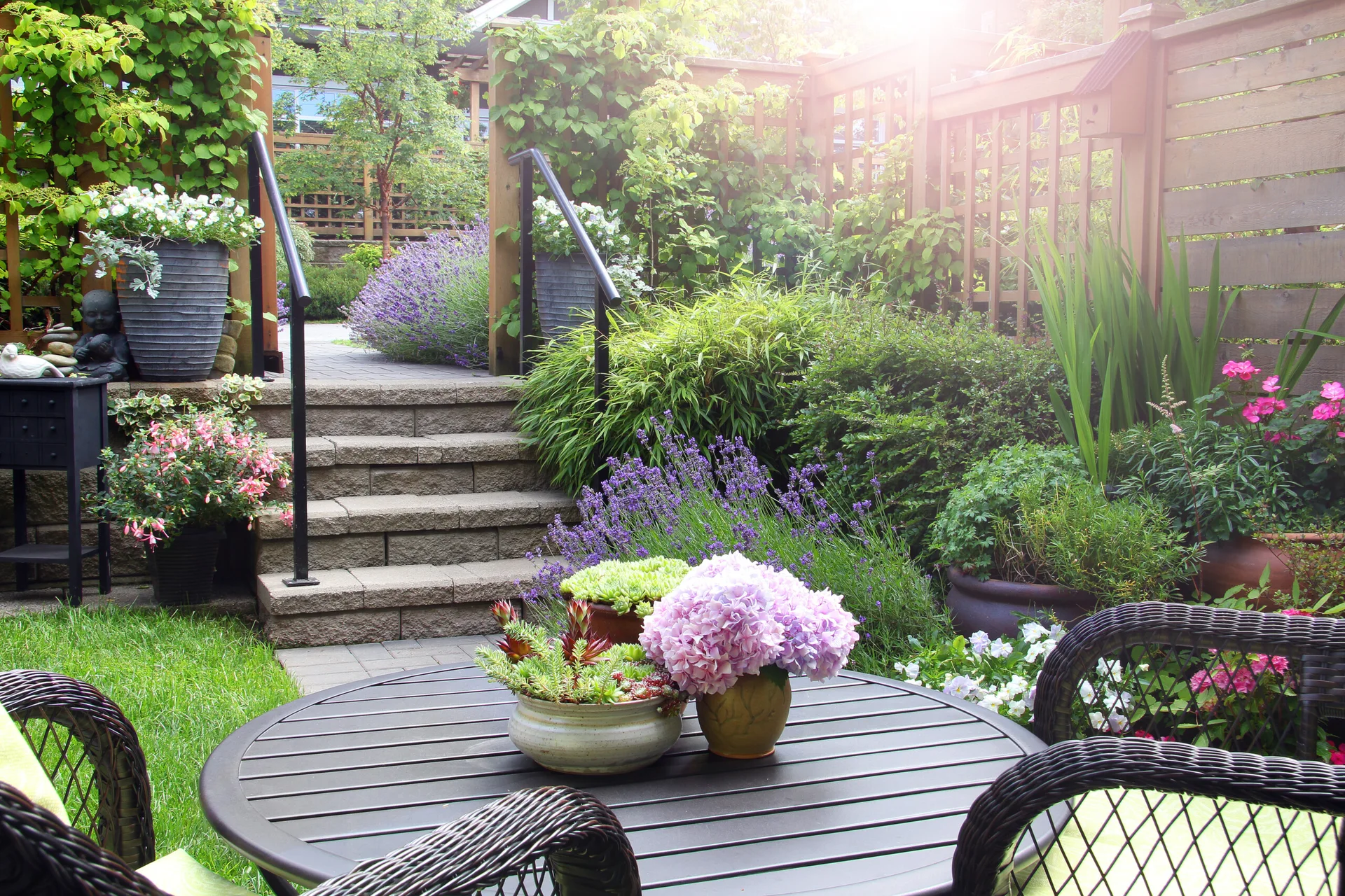A black metal dining table with pink hydrangeas sits near stone steps in a lush cottage garden surrounded by purple flowers and wood fencing.