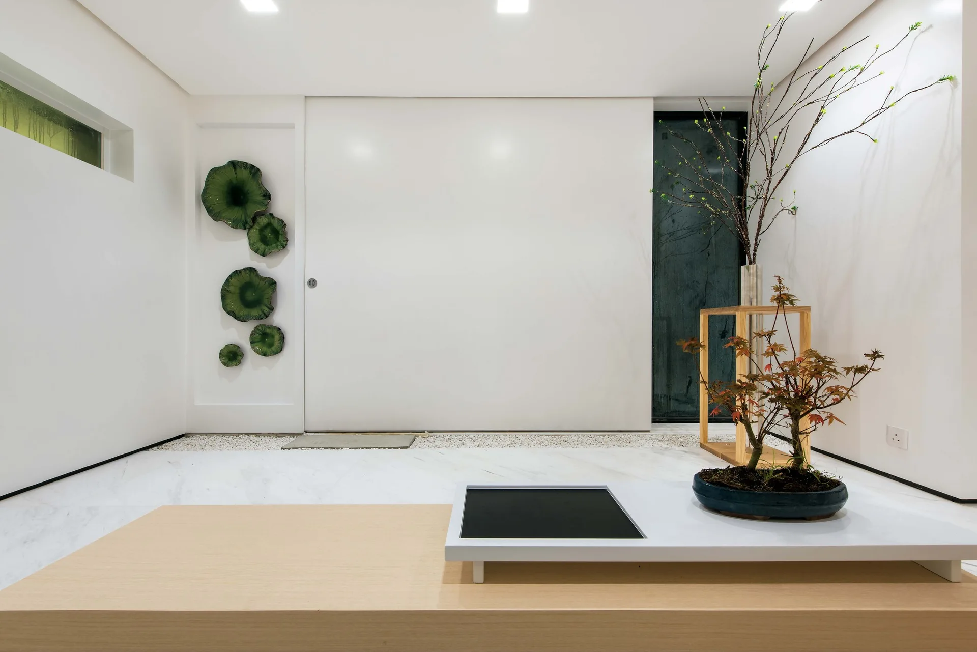 A minimalist white foyer featuring a low wooden platform with a bonsai plant, a large sliding panel door, and green ceramic disc wall decorations.