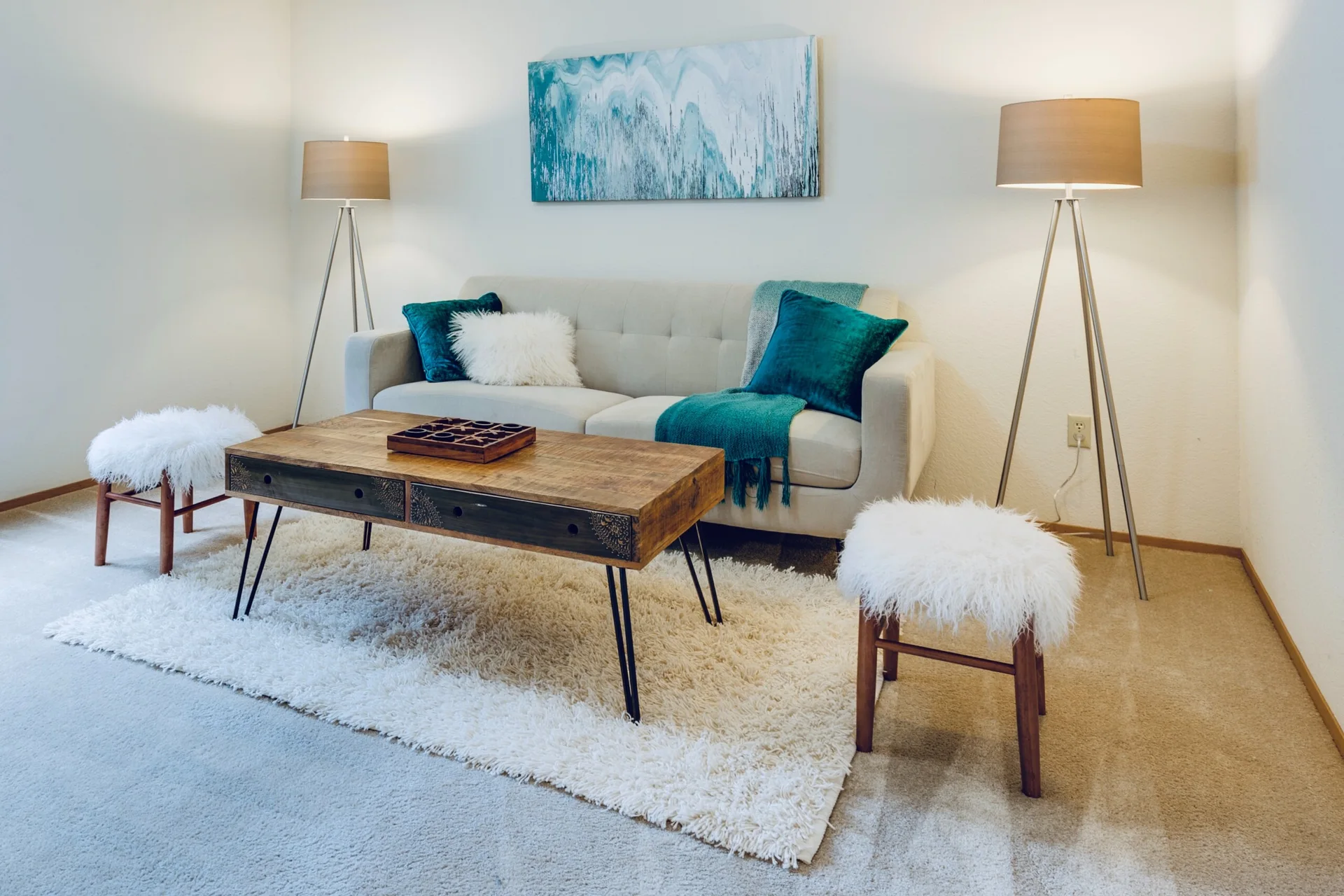 A beige sofa sits between two tripod floor lamps on a shag rug, accompanied by a wooden coffee table and two white furry stools.