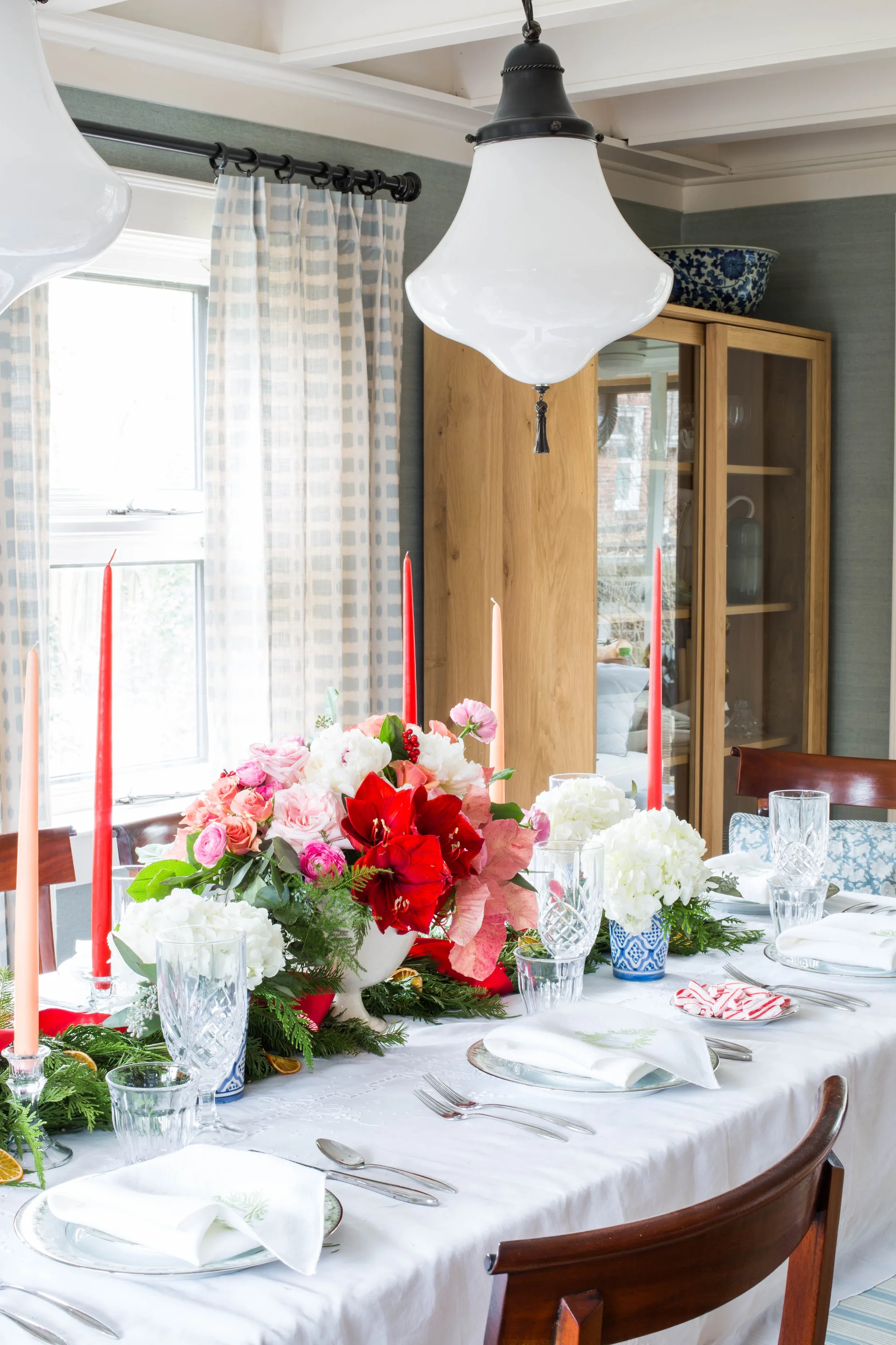 A formal dining table is set with white linens, crystal glassware, red taper candles, and a festive floral centerpiece.