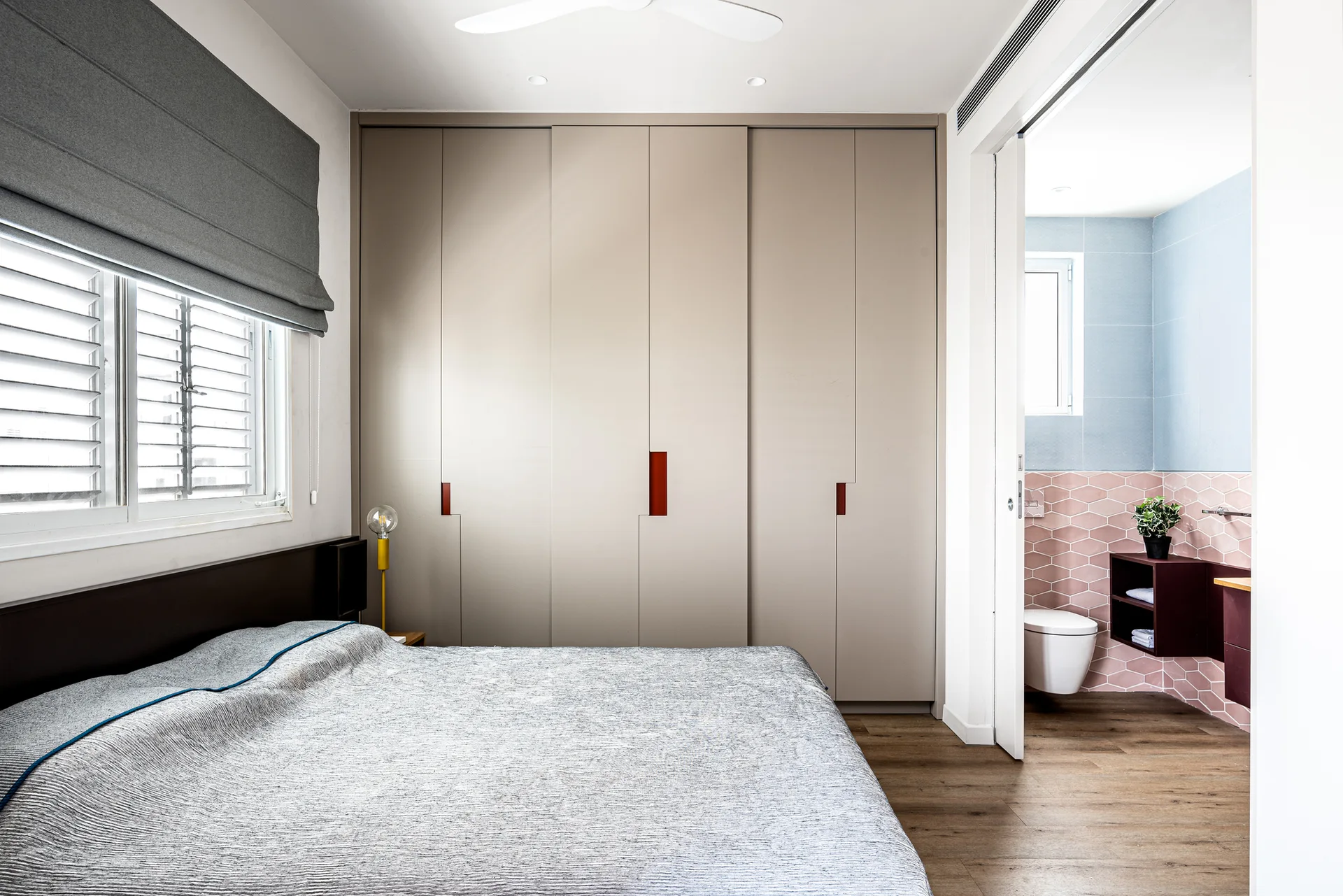 A modern bedroom features a large built-in tan wardrobe, a low-profile bed, and an open sliding door revealing a bathroom with pink hexagonal tiles.
