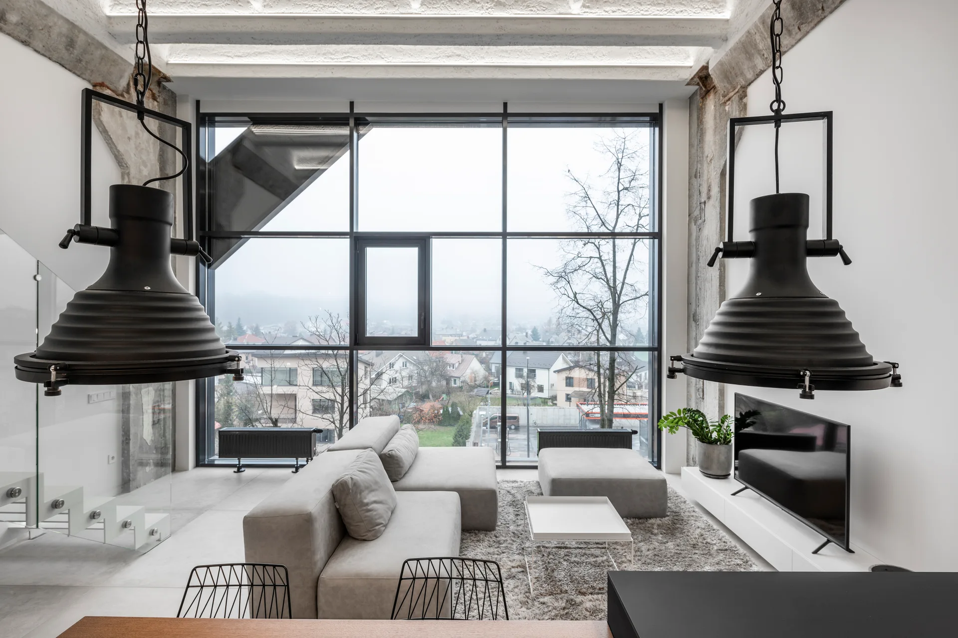 An industrial style living room features a grey modular sofa, large grid window, and oversized black pendant lights hanging from concrete beams.