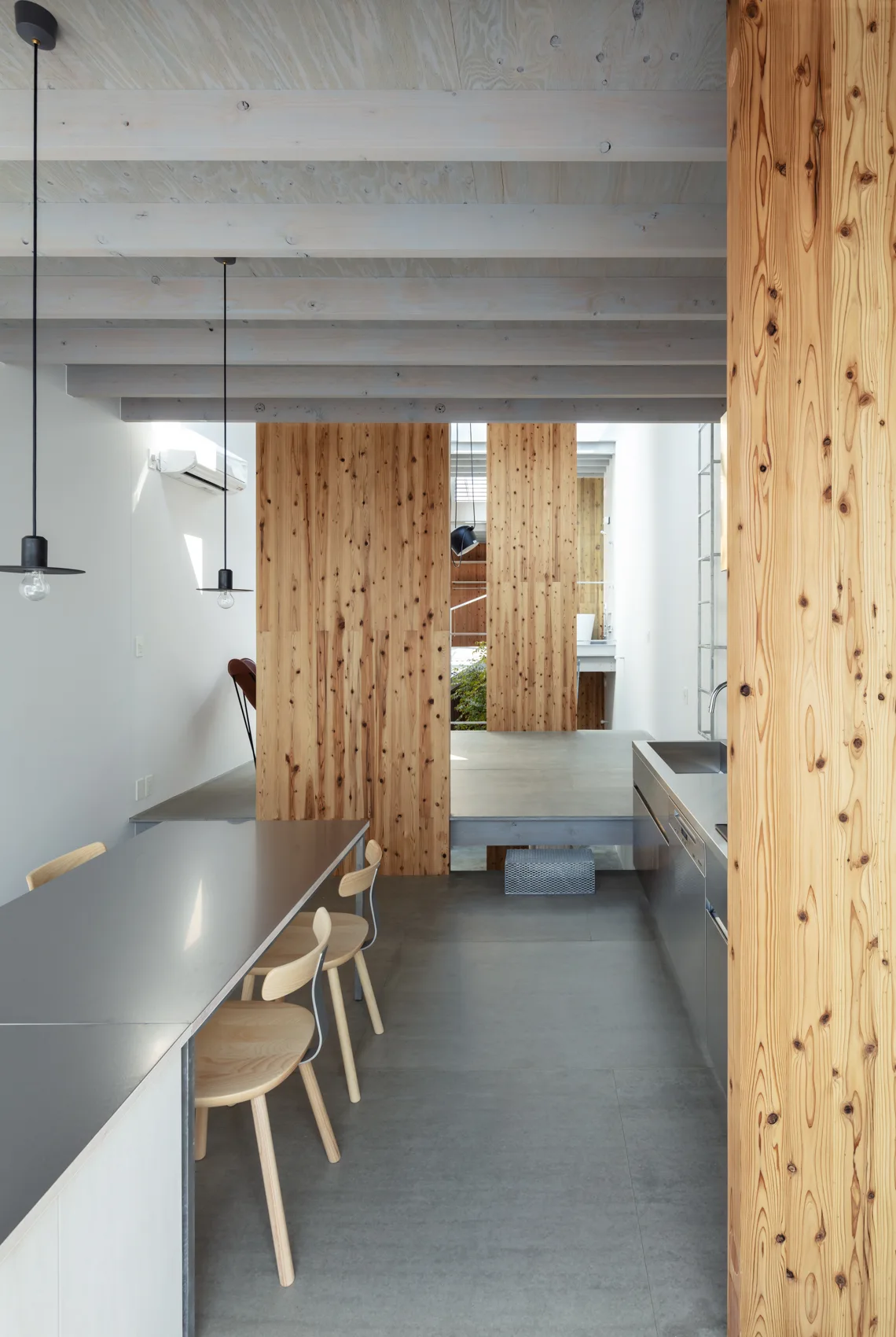 A minimalist kitchen and dining area featuring a grey dining table with wood chairs, a stainless steel counter, and tall knotty wood partitions.