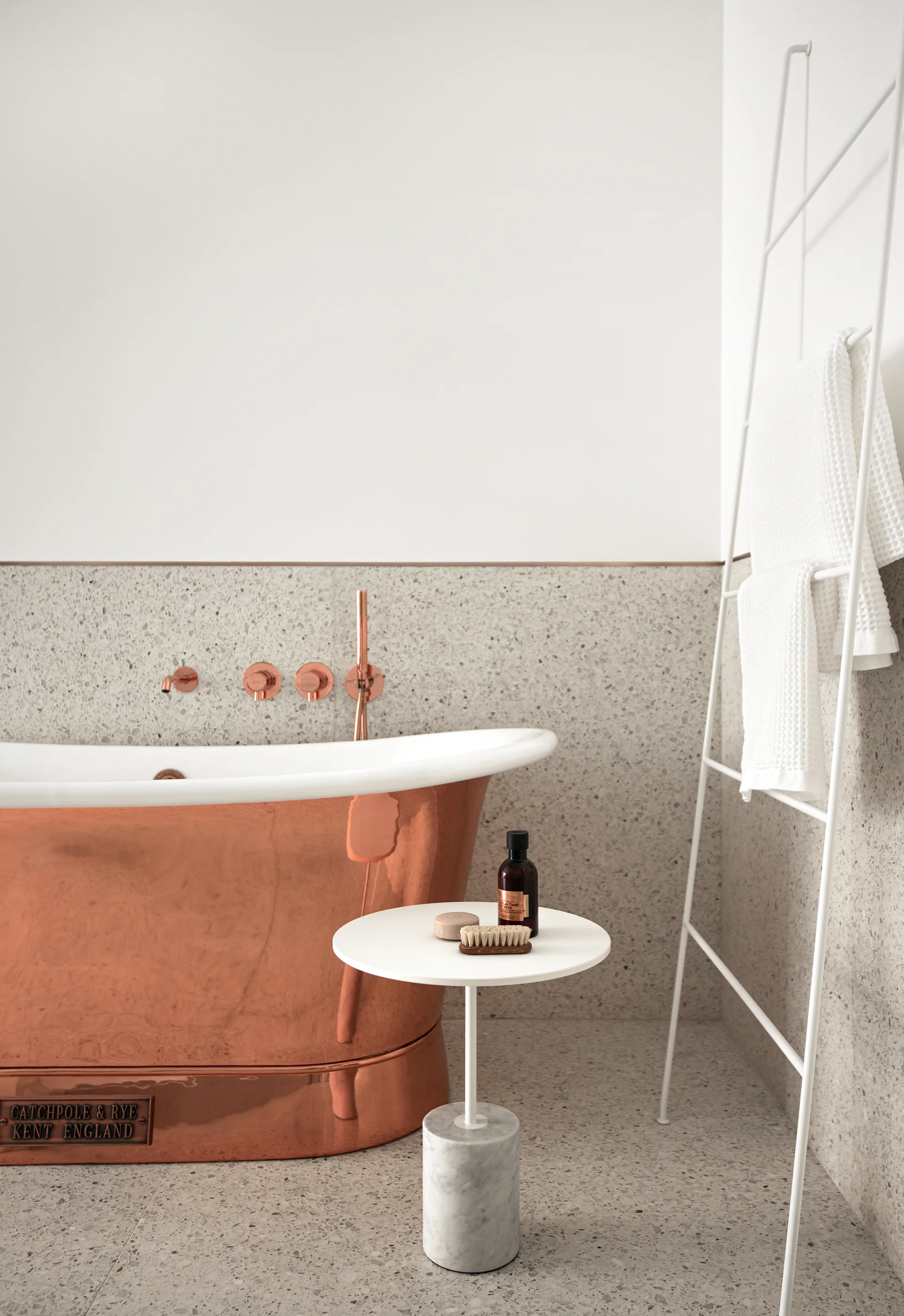 A modern bathroom features a freestanding copper bathtub set against a grey terrazzo tiled wall next to a white metal towel ladder.