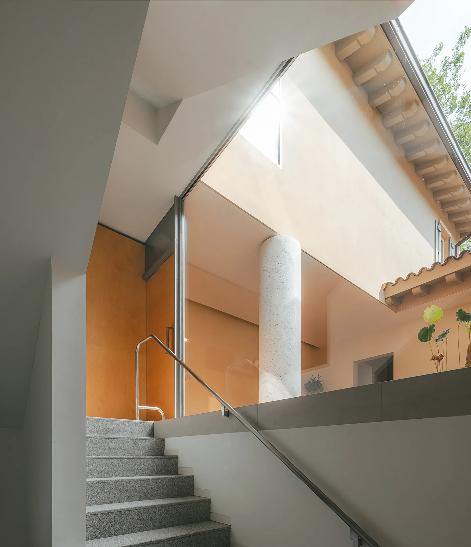 A modern grey stone staircase with a metal railing rises next to a tall glass wall looking out to a courtyard with a white column.