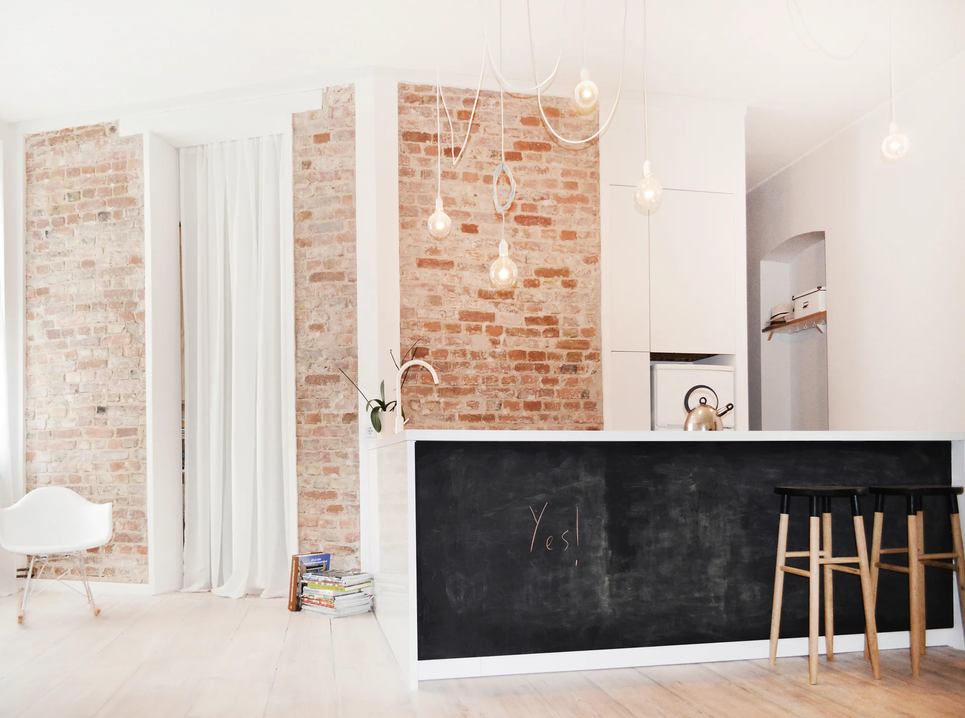 A modern kitchen with a chalkboard-fronted island, wooden stools, exposed brick walls, and hanging bulb pendant lights.