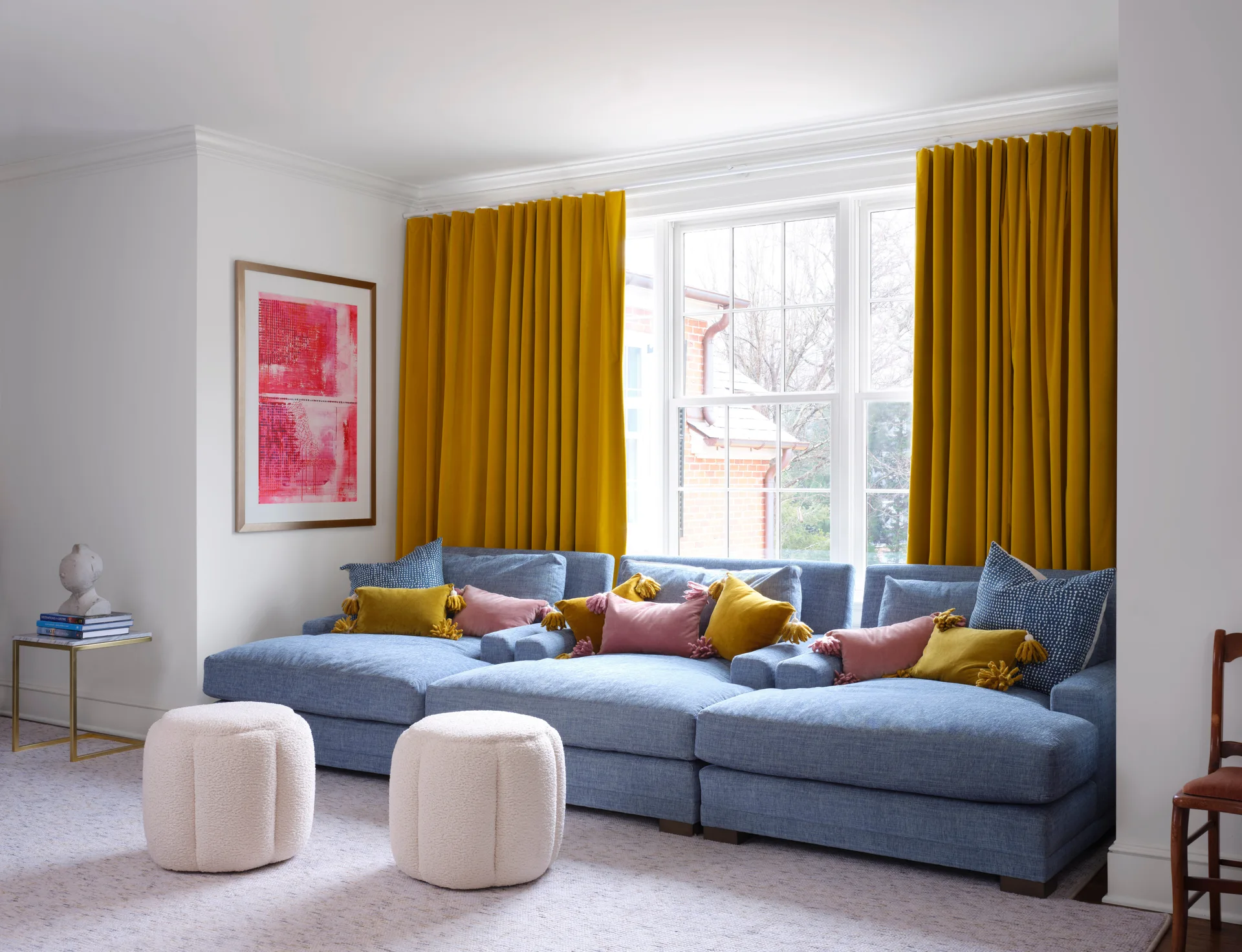 A blue modular sectional sofa is accented with yellow and pink pillows beneath bright yellow curtains in a white living room with two white round ottomans.