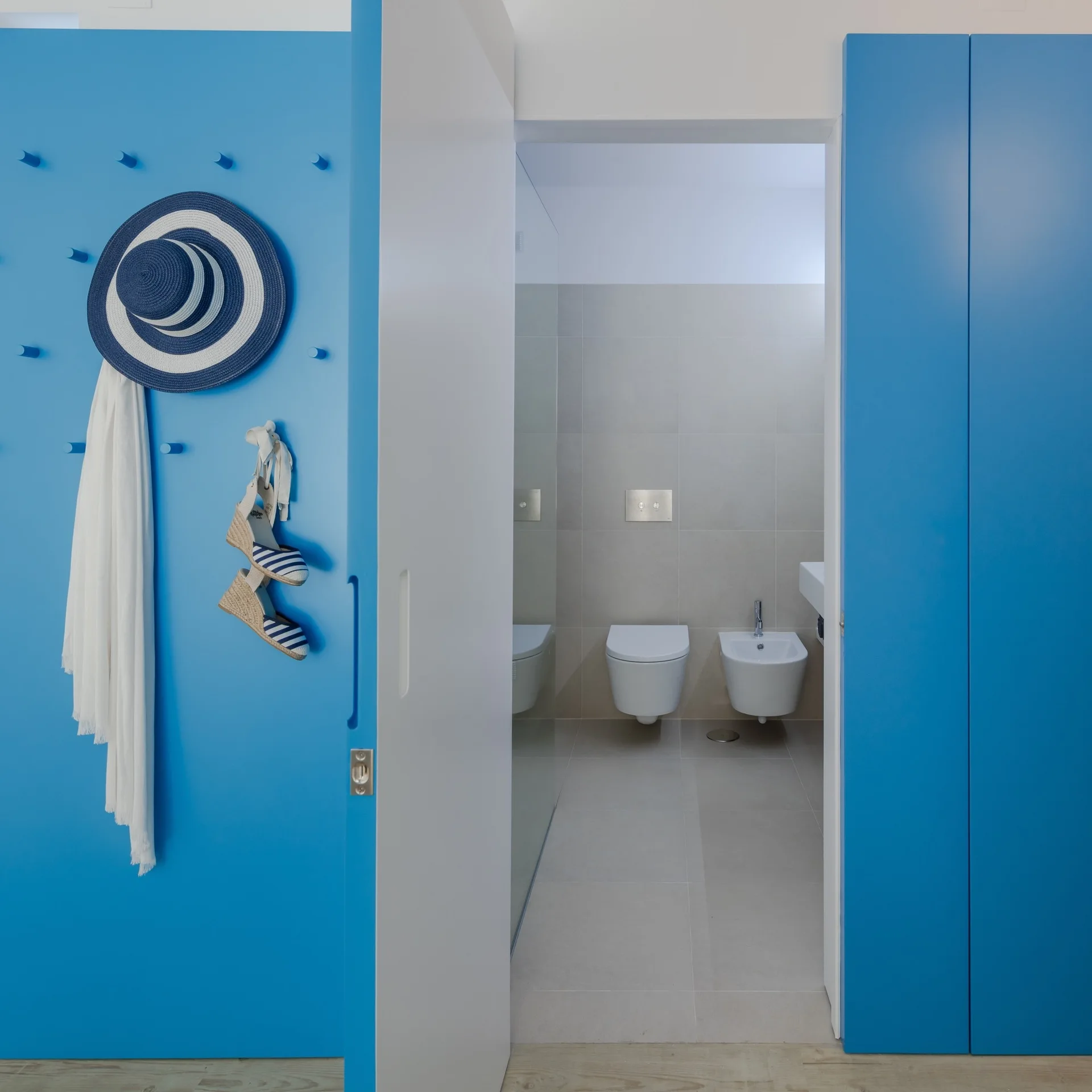 A view through a doorway into a modern bathroom with a wall-mounted toilet, contrasted by a bright blue paneled storage wall with matching pegs.