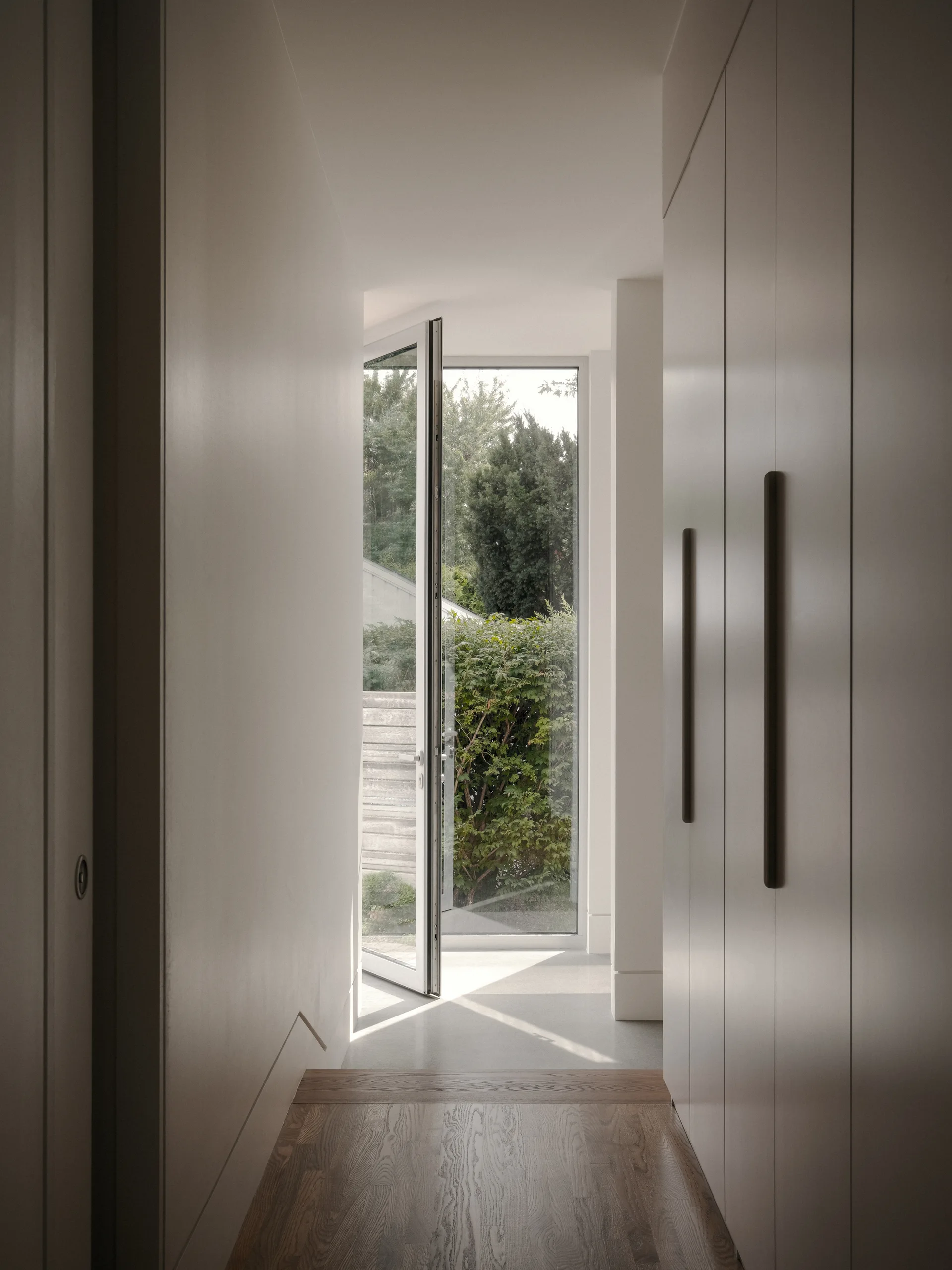 A minimalist hallway features tall built-in cabinets with dark vertical handles, wood flooring, and a glass door opening to a garden.