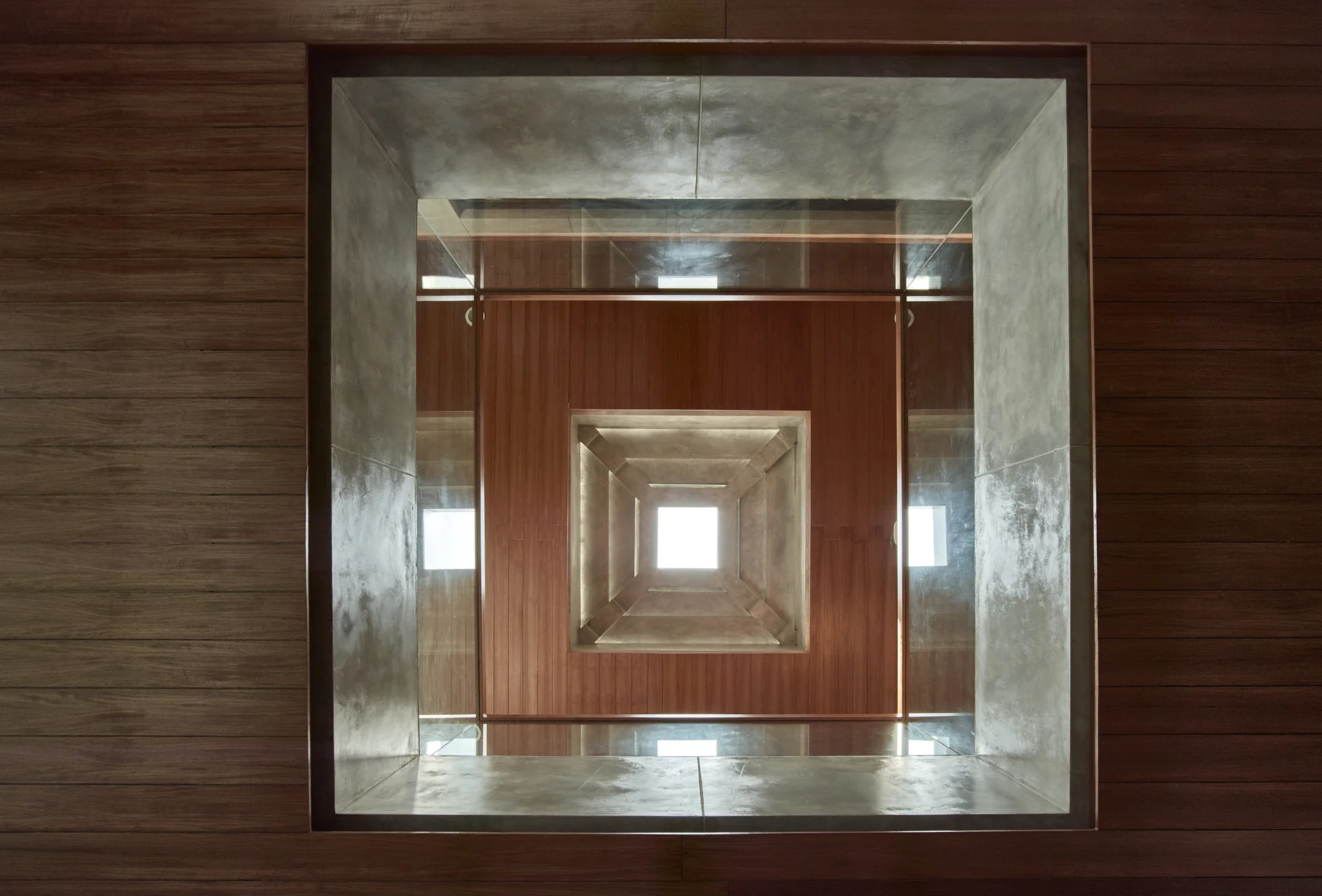 A low-angle view of a geometric, recessed square skylight framed by alternating layers of warm wood paneling and smooth grey concrete.