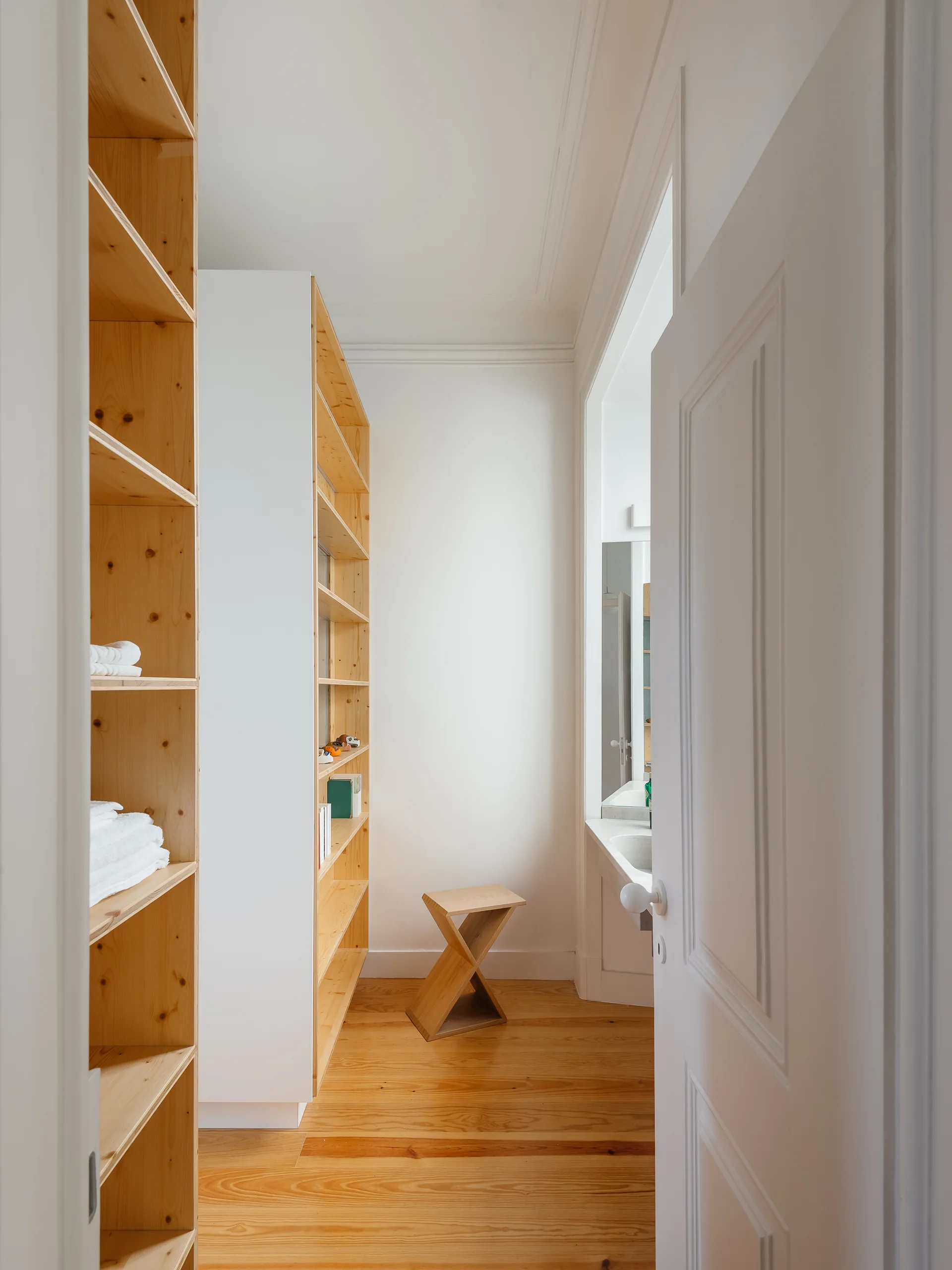 A bright walk-in closet features open built-in pine shelving units, hardwood floors, a white paneled door, and a small wooden x-base stool.