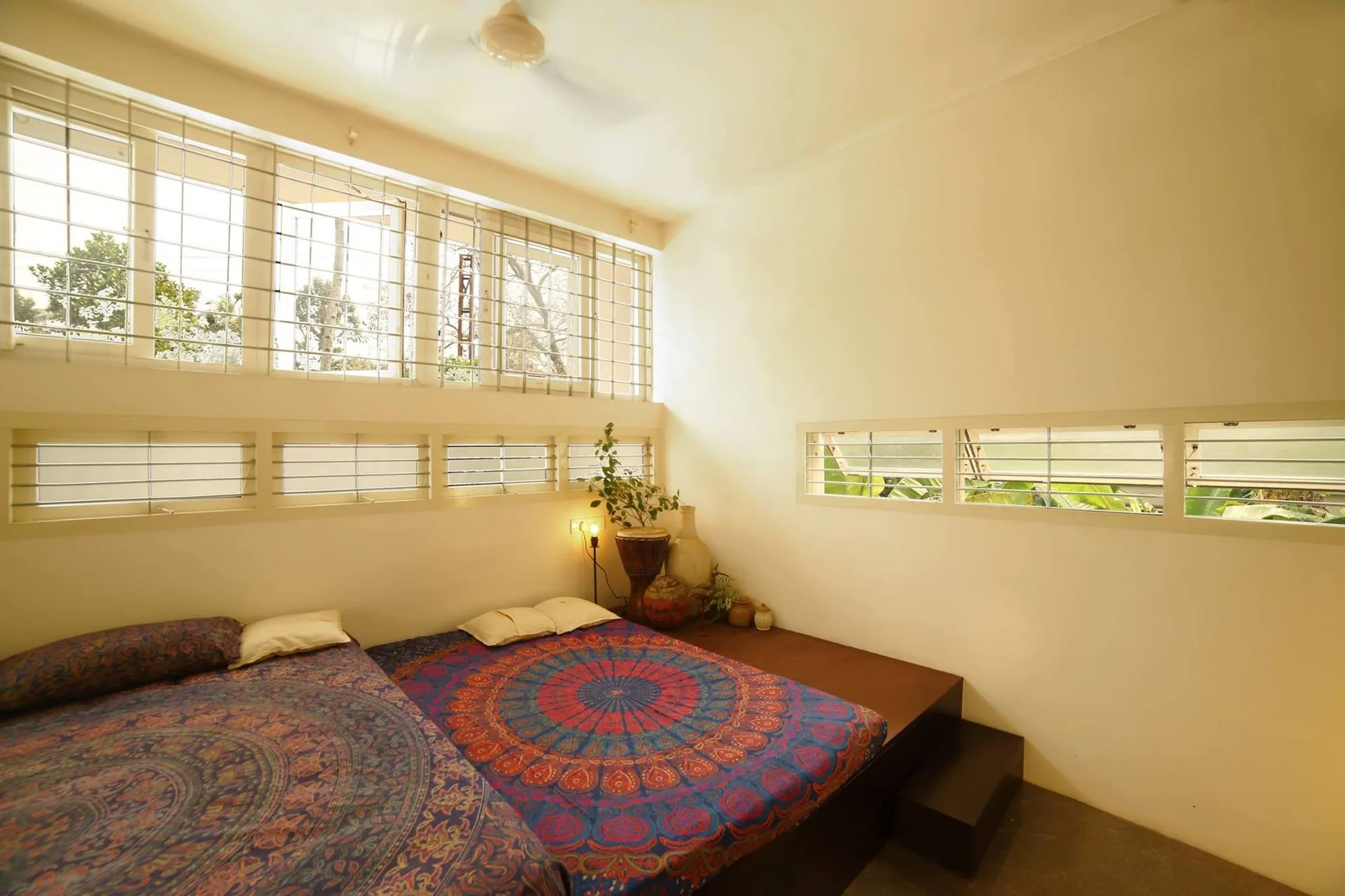 A minimalist bedroom features a low dark wood platform bed with colorful mandala-print bedding beneath large white windows with metal grilles.