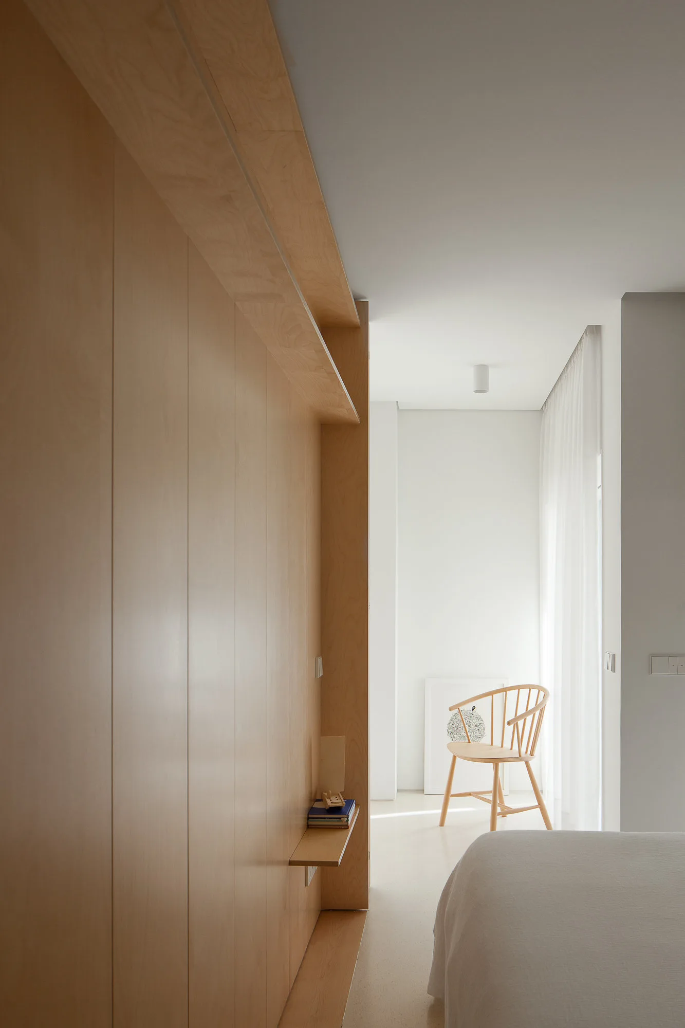 A minimalist bedroom featuring light wood built-in cabinetry, a wooden spindle chair by a window with sheer white curtains, and the edge of a bed.