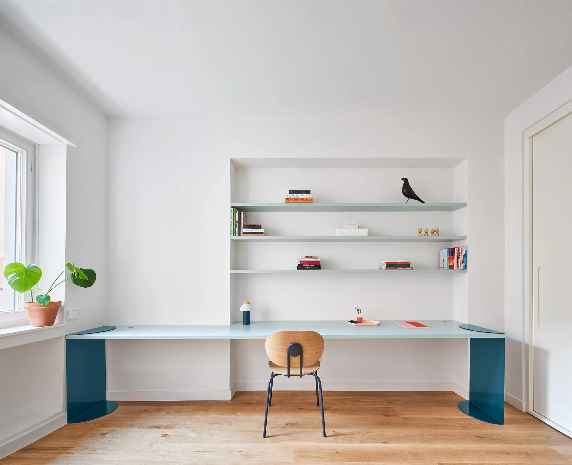 A minimalist home office features a long pale-blue desk supported by curved teal metal legs under three matching recessed wall shelves.
