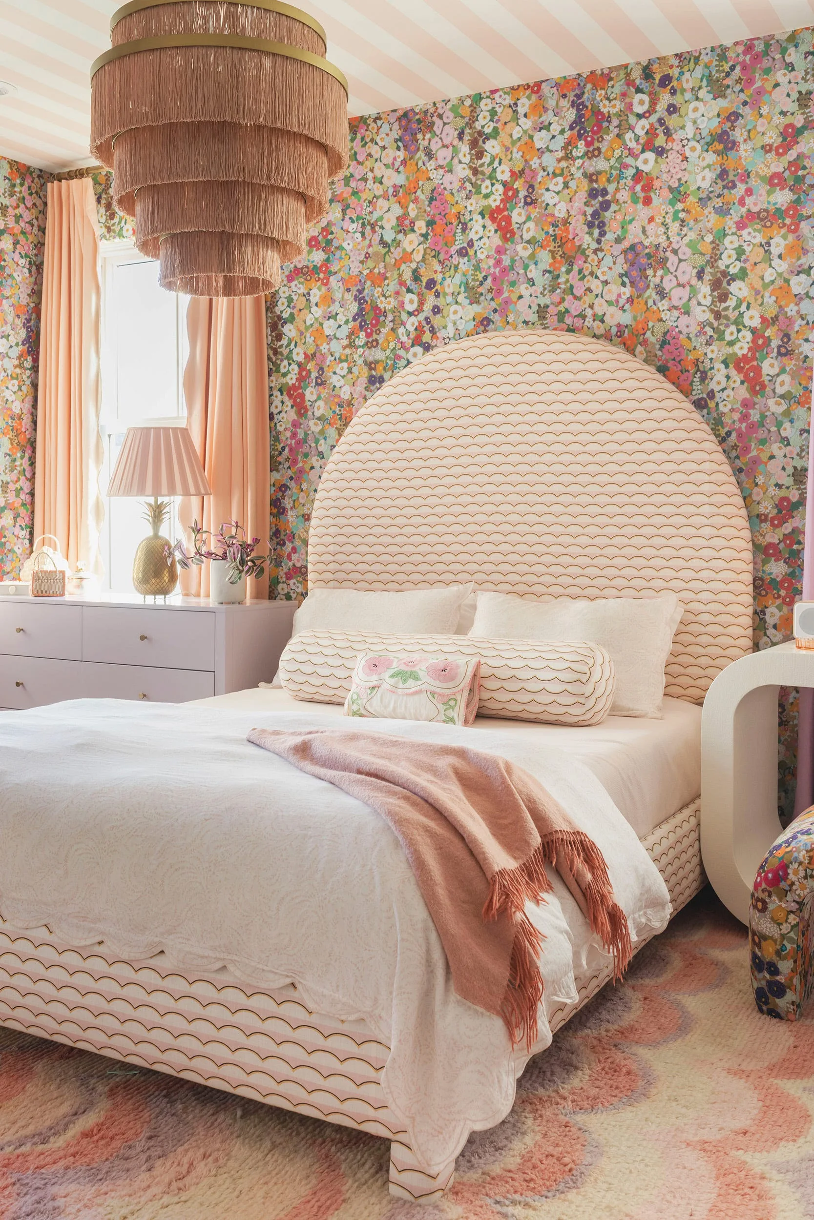 A whimsical bedroom features a large arched headboard with a scalloped pattern, dense floral wallpaper, a lavender dresser, and a tiered fringe chandelier.