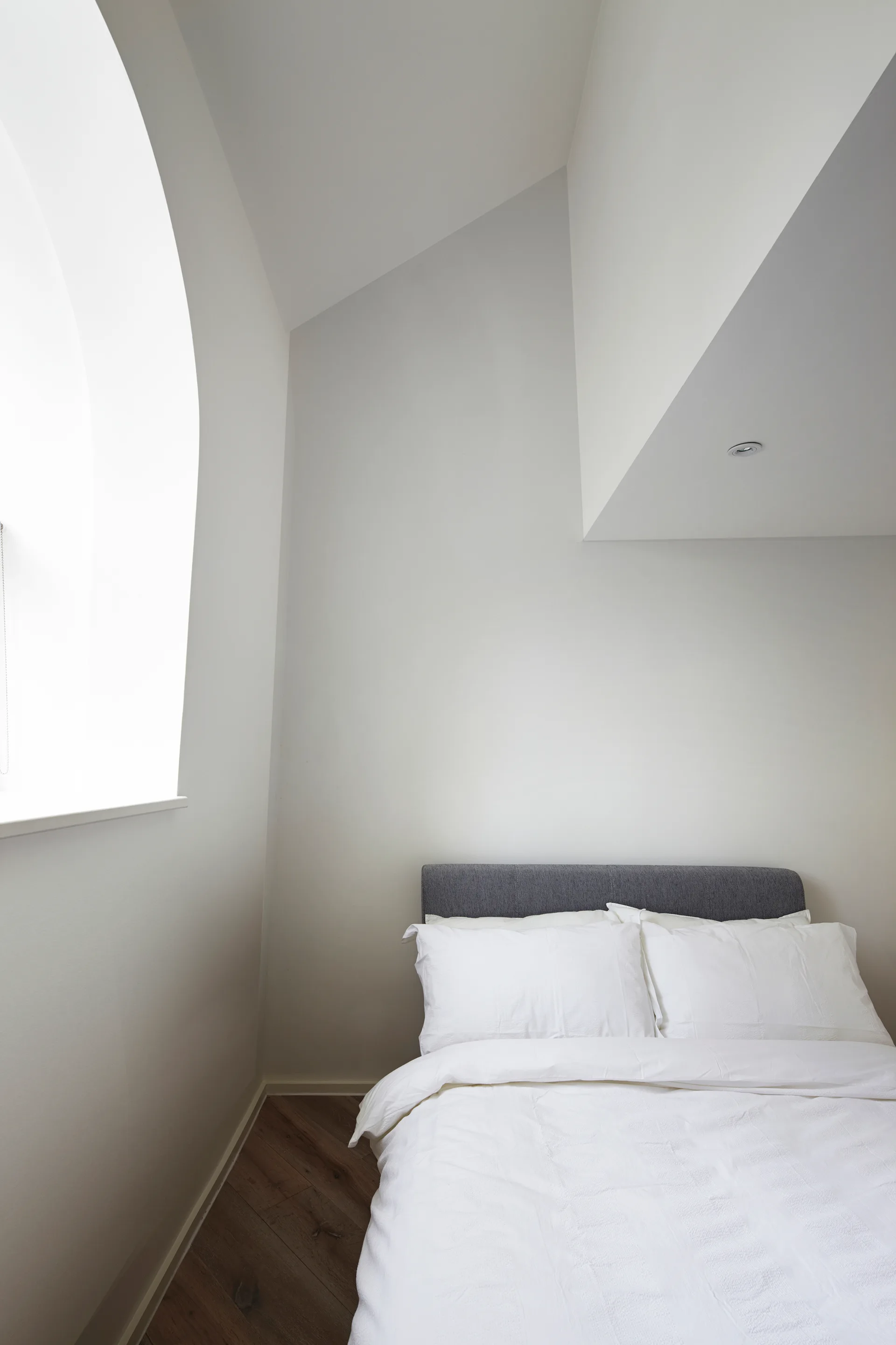 A minimalist bedroom features a charcoal grey upholstered bed headboard against white walls beneath a sloped ceiling and next to a deep arched window.