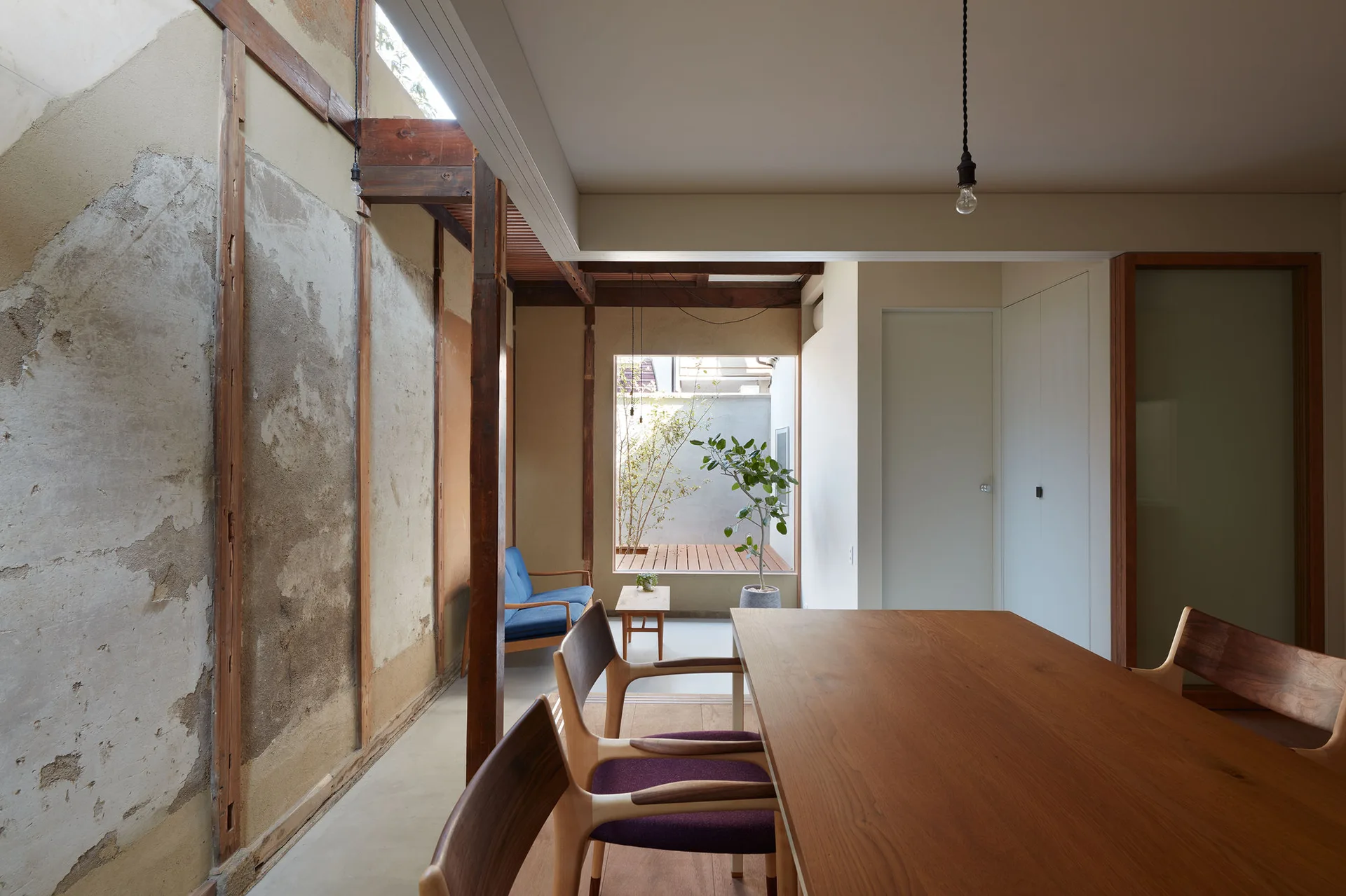 A minimalist dining room features a long wooden table, chairs with purple cushions, an exposed plaster wall, and a view of a small rear courtyard.