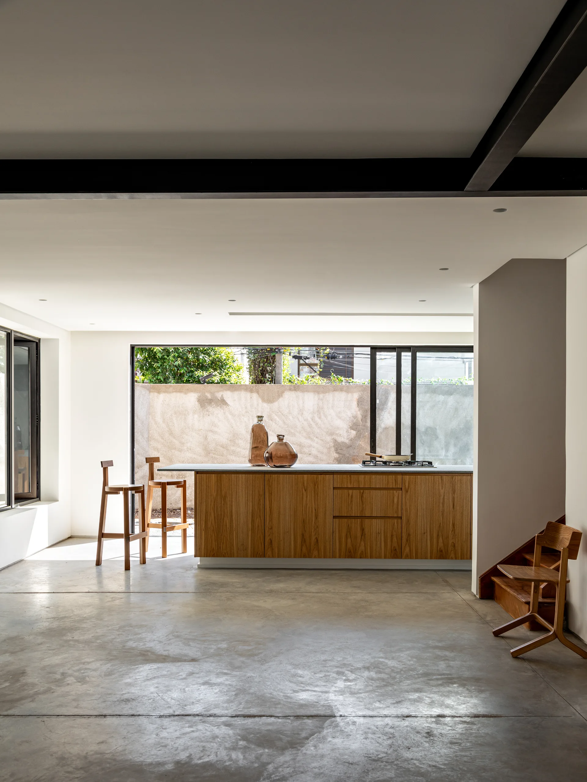 A minimalist kitchen featuring a wood-fronted island with bar stools, polished concrete floors, and a large sliding glass patio door.
