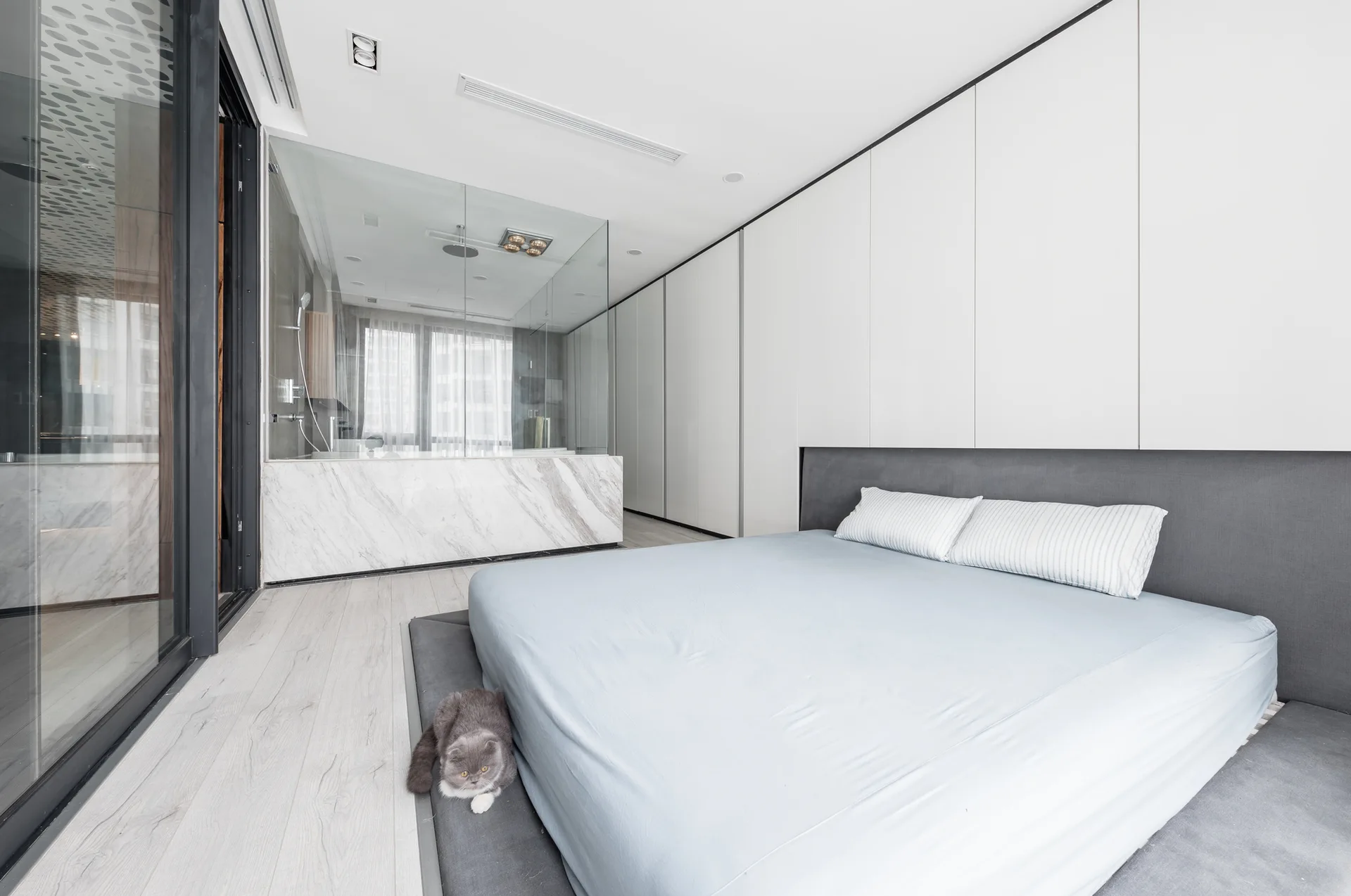 A minimalist bedroom features a low-profile blue bed, floor-to-ceiling white cabinetry, and a glass-enclosed bathroom on a marble base.