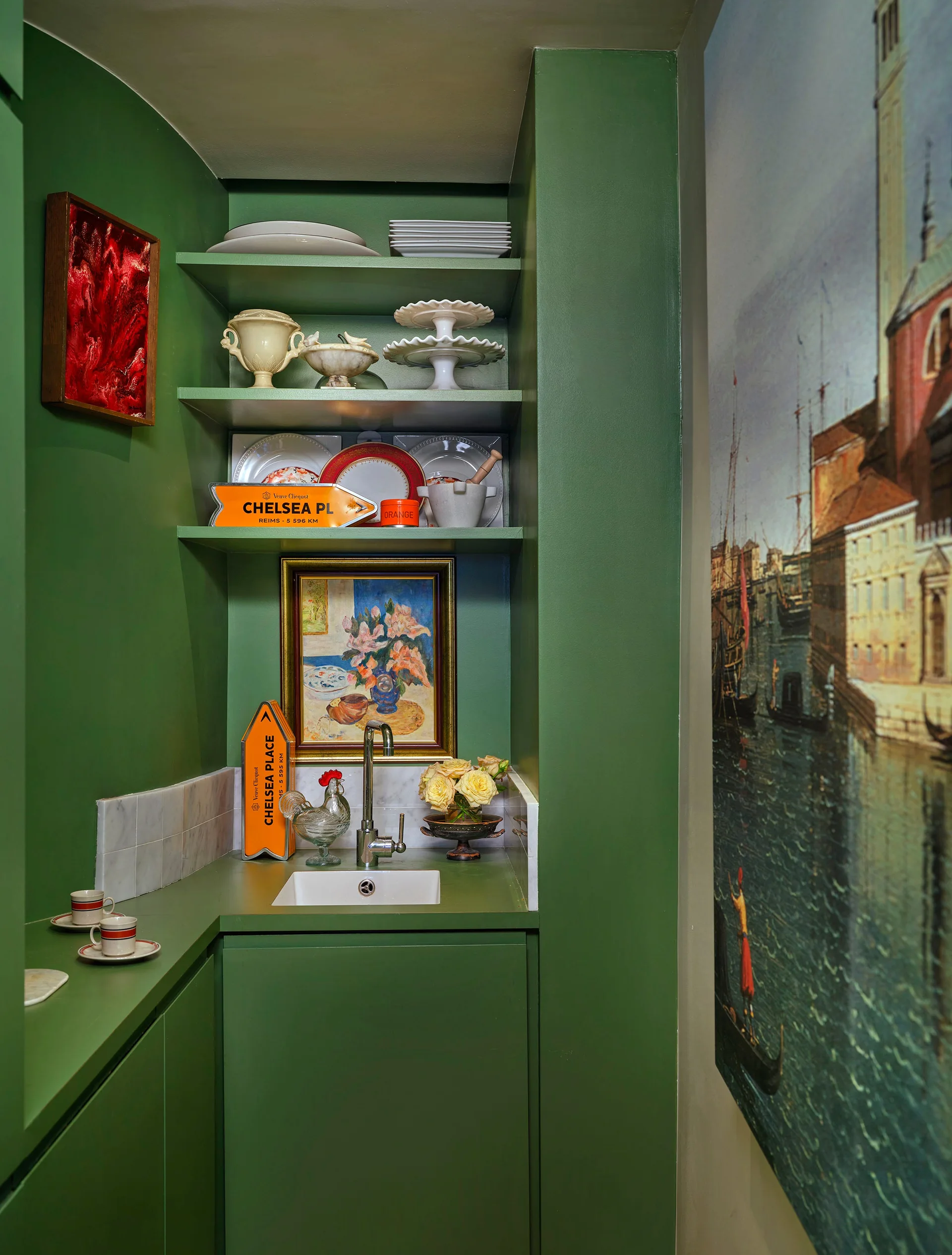 A small green wet bar features a recessed sink with a chrome faucet, built-in shelves displaying plates and artwork, and a nearby wall mural.