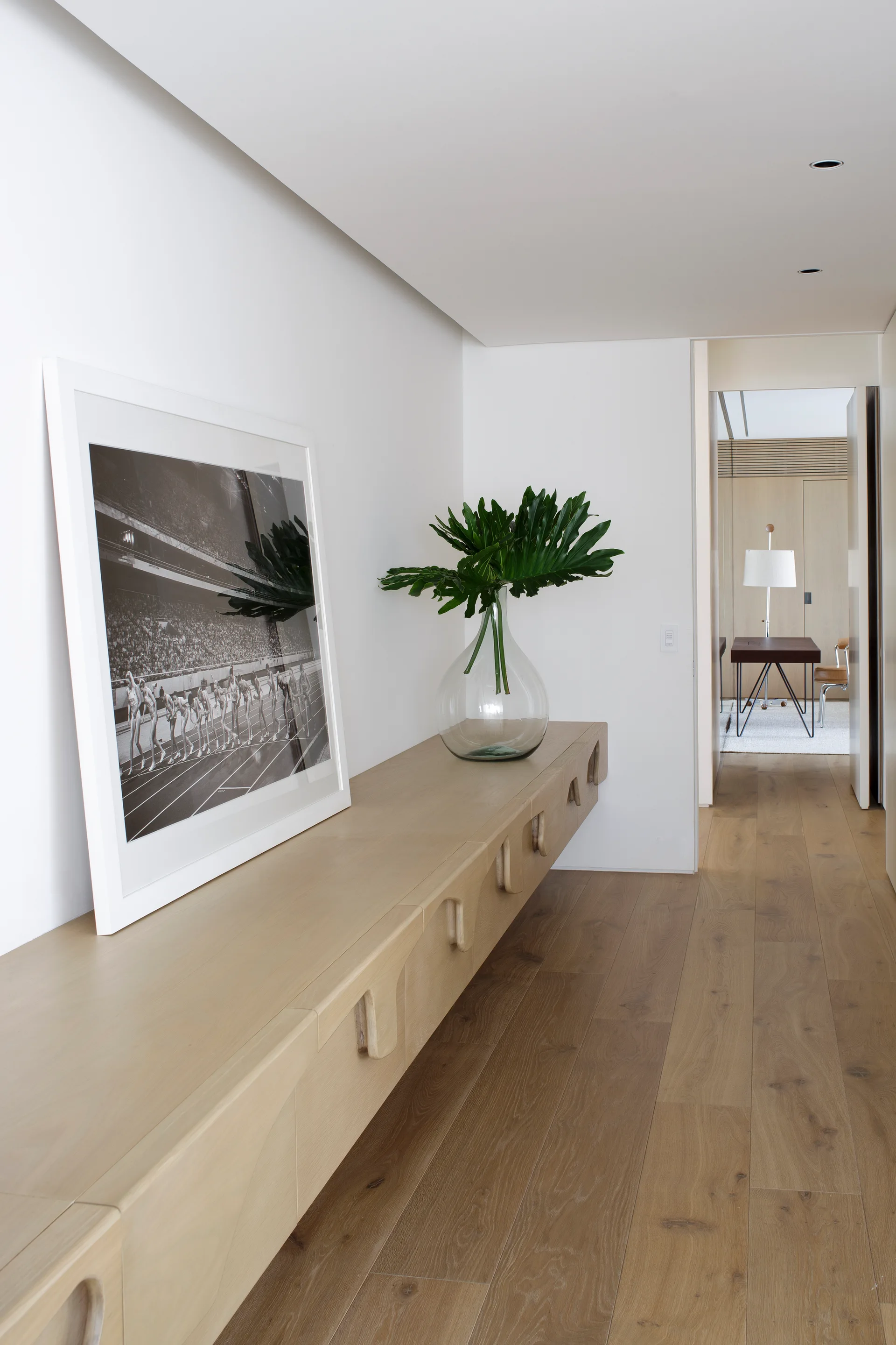 A minimalist white hallway features a floating light wood console styled with a large black and white framed print and a clear glass vase with green leaves.