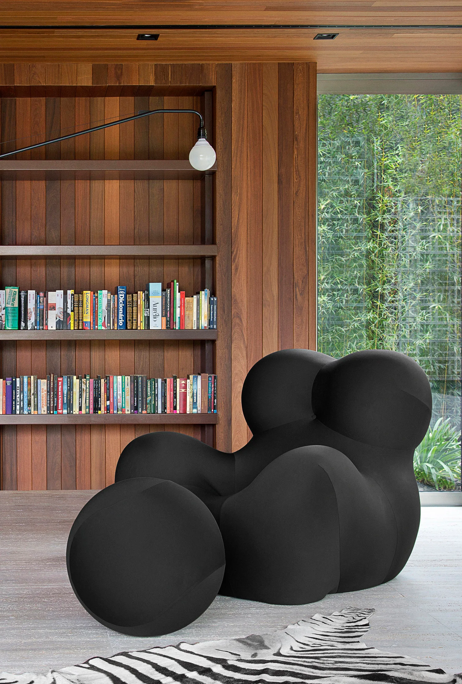 A sculptural black armchair sits on a zebra stripe rug next to a built-in wooden bookshelf and a large window.