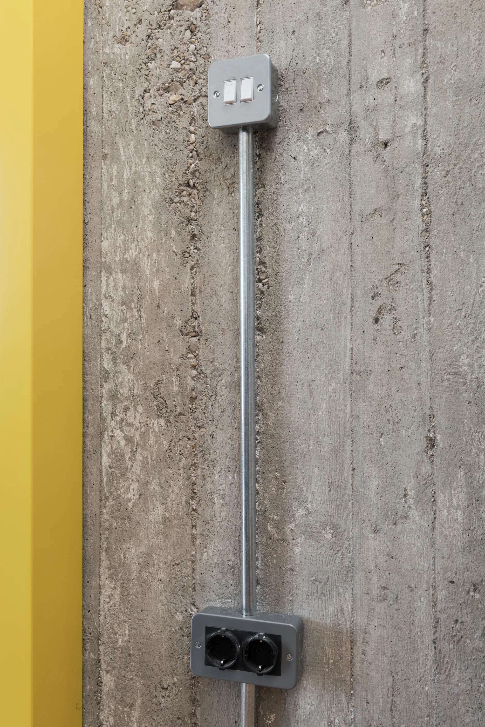 A metal light switch and power outlet with exposed metal conduit mounted on a rough concrete wall next to a yellow painted vertical panel.