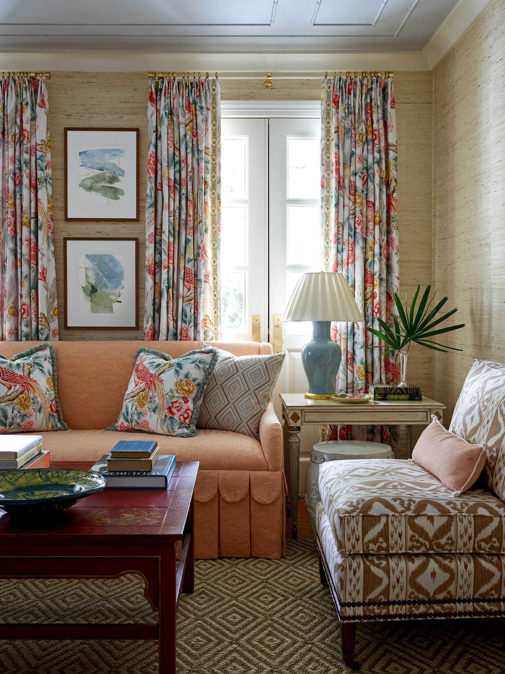 A traditional living room seating area features a peach sofa, a patterned ikat armchair, a red coffee table, and floral curtains framing a glass French door.