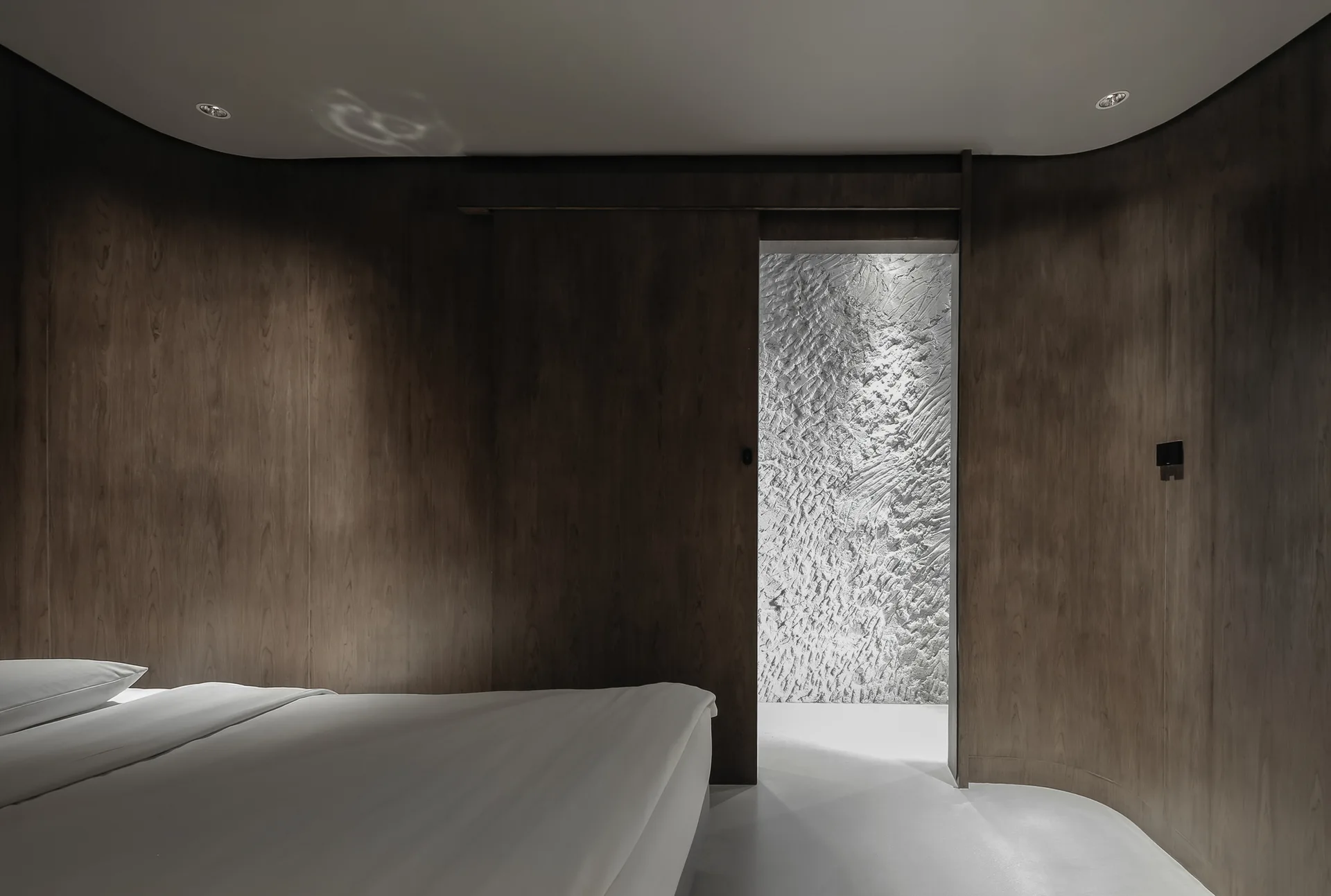 A minimalist bedroom features curved dark wood paneled walls, a white bed, and a sliding door that opens to a highly textured white wall.