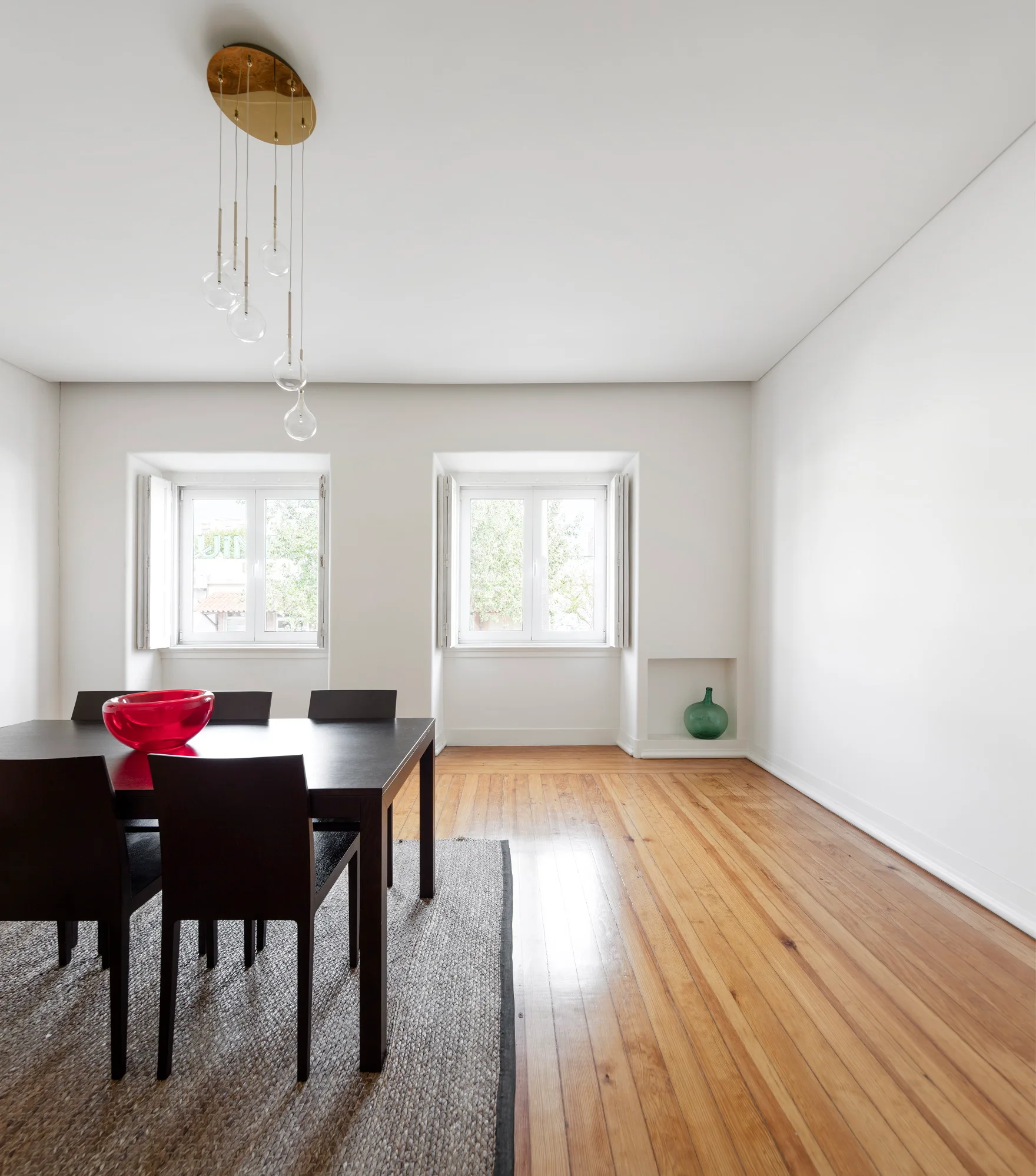 A minimalist dining room features a black table and chairs on a textured rug, wooden floors, white walls, and a multi-globe pendant light.