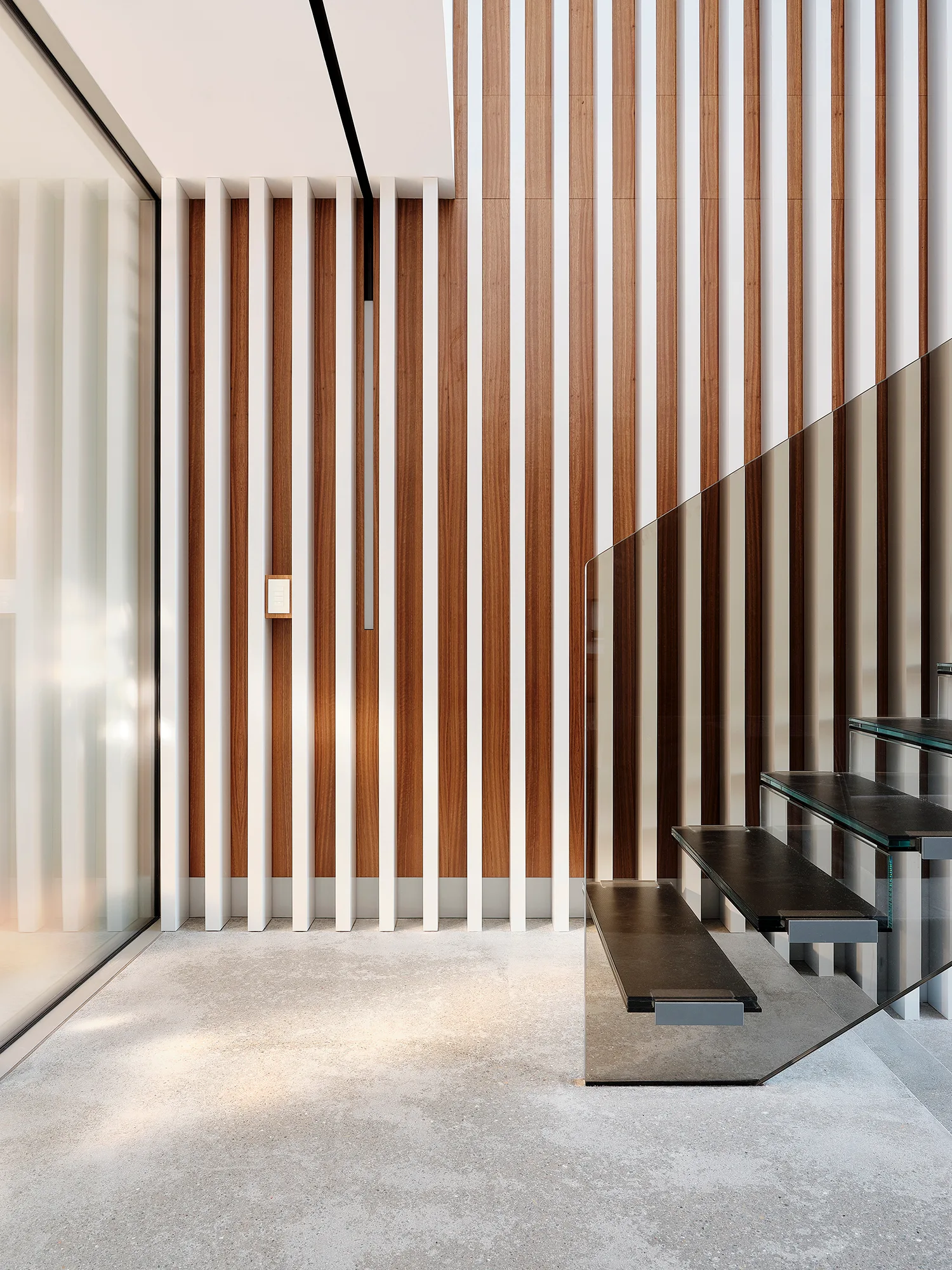 A minimalist staircase with dark steps and a tinted glass railing stands next to a tall wall made of alternating white and wood-toned vertical slats.