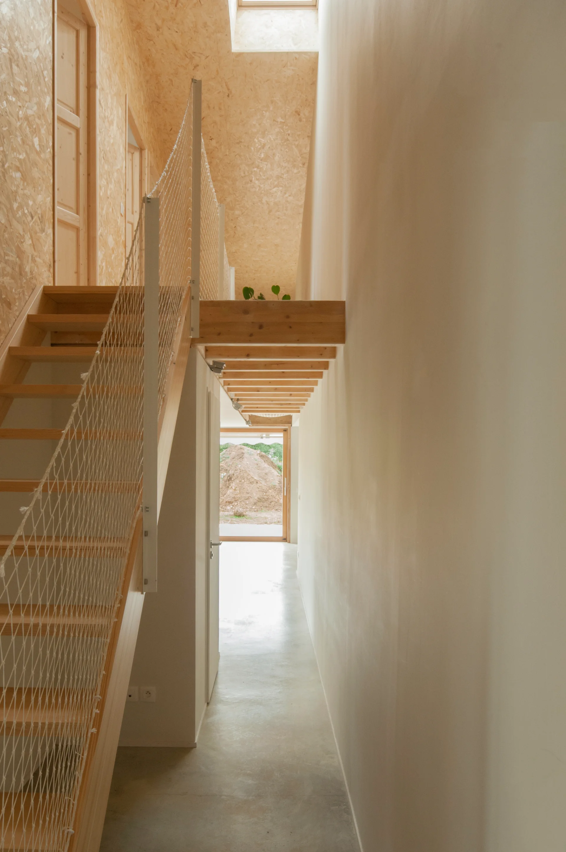 A wooden staircase with a white net railing ascends in a minimalist hallway with concrete floors and oriented strand board walls.