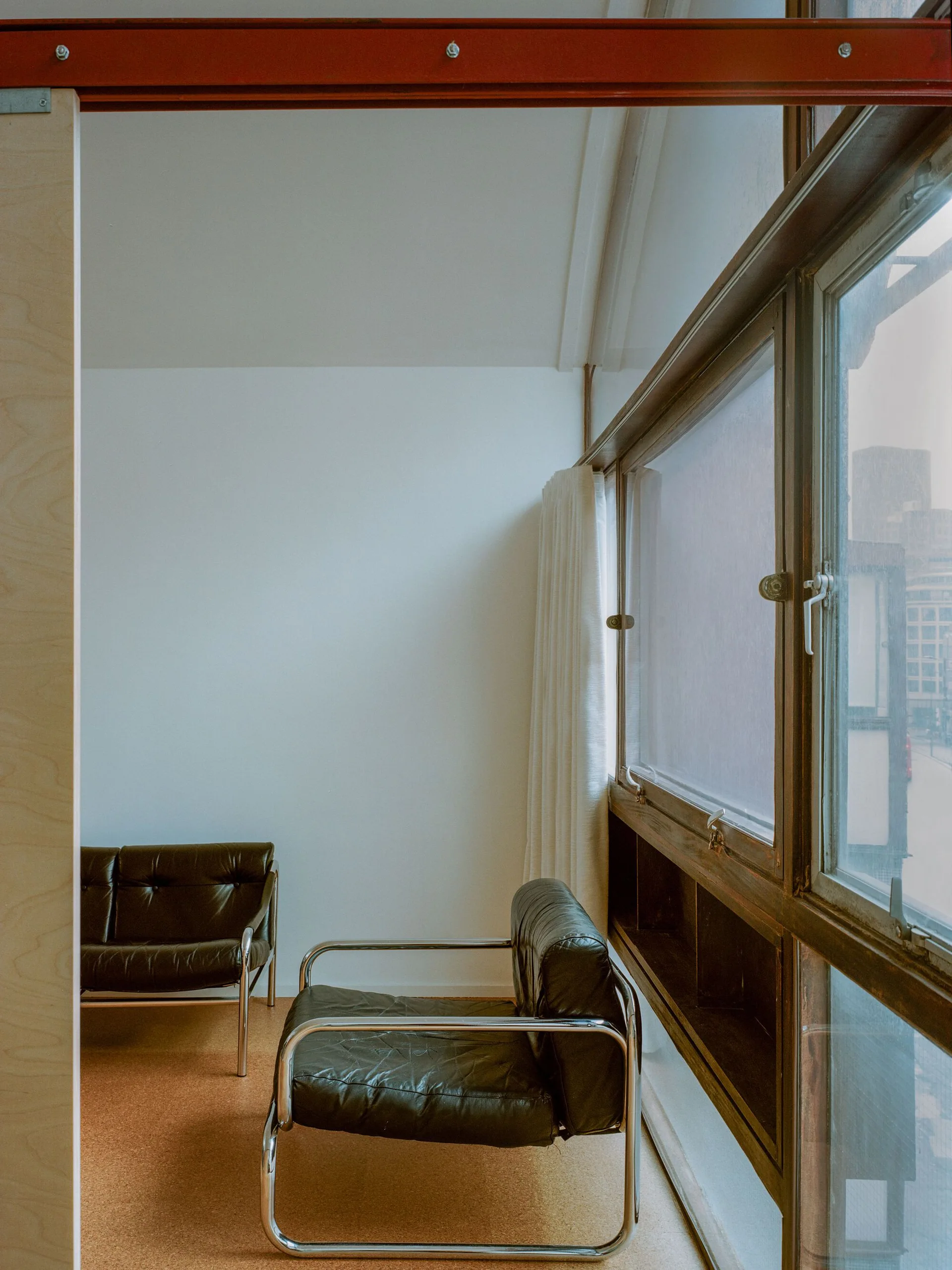 A modernist living space features a black leather armchair with a chrome frame set beside a large wood-framed window.