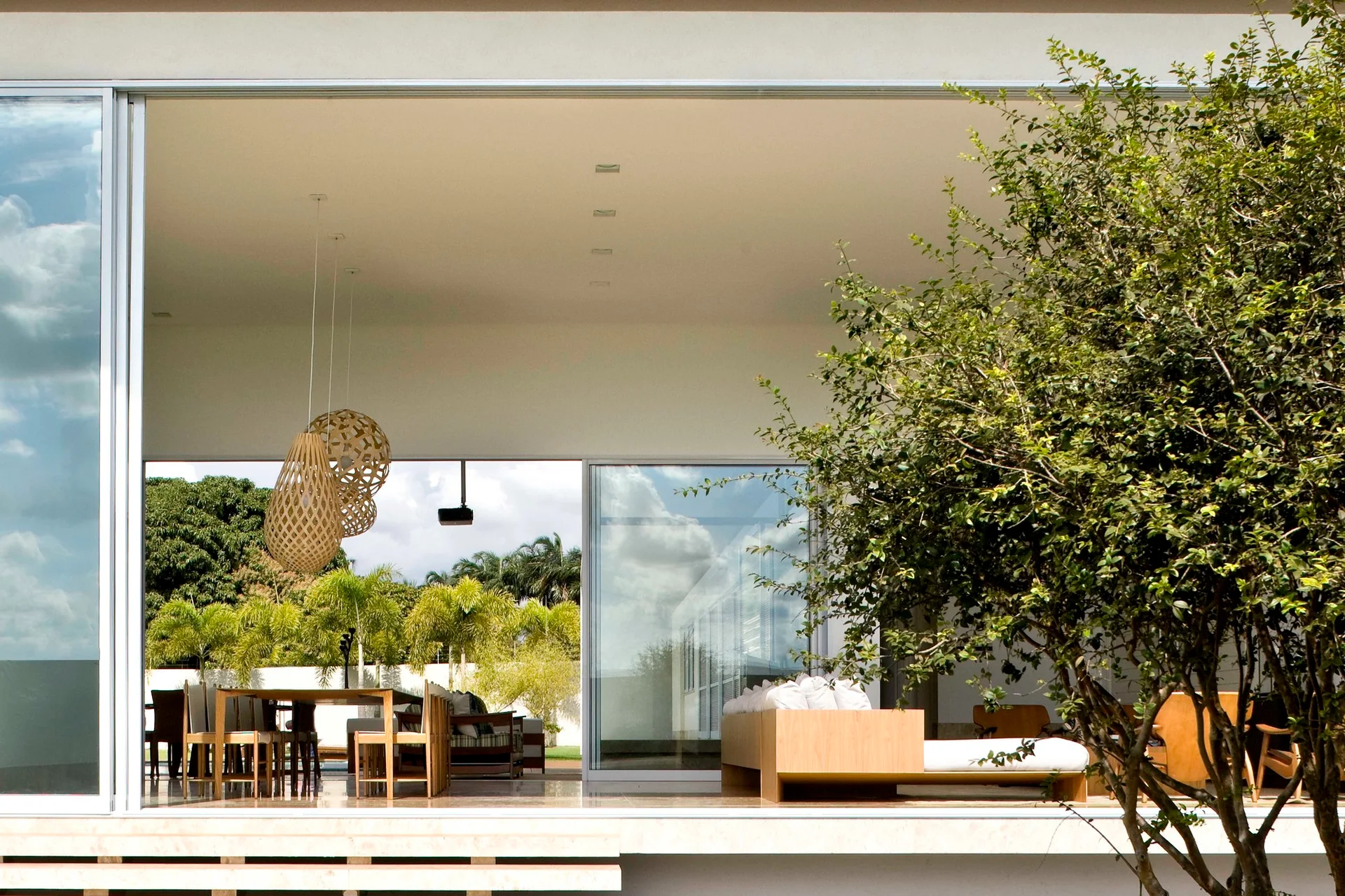 A covered minimalist patio features a wooden dining table with woven basket pendant lights and a low-profile sofa framed by large glass doors.