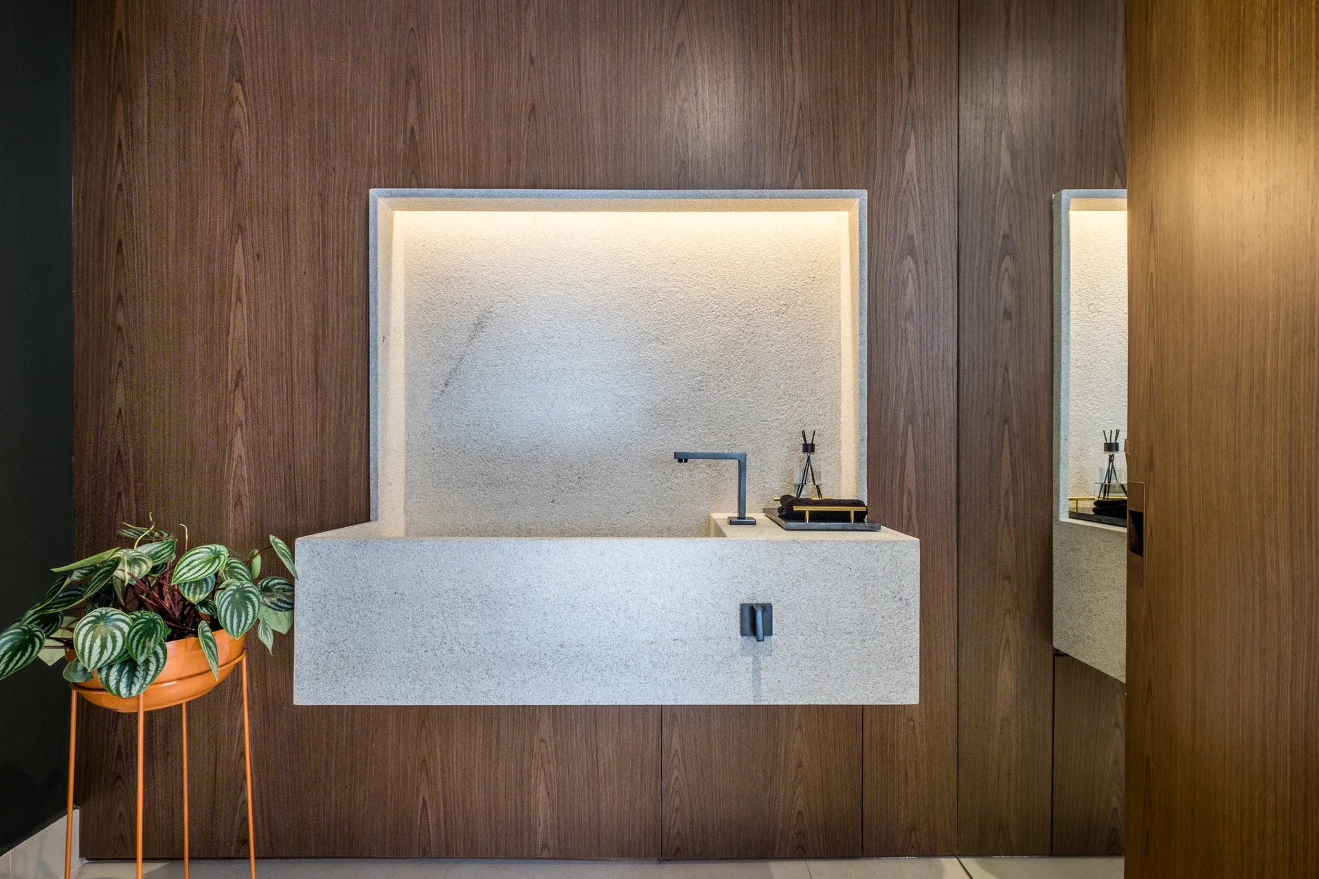 A wall-mounted grey stone washbasin set into a lighted niche against a dark wood paneled wall with a small plant on the left.