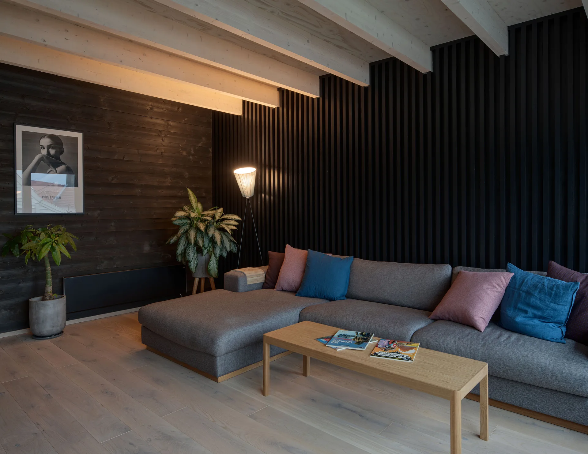 A grey sectional sofa sits against a black vertical wood slat wall under exposed light wood ceiling beams in a modern living room.