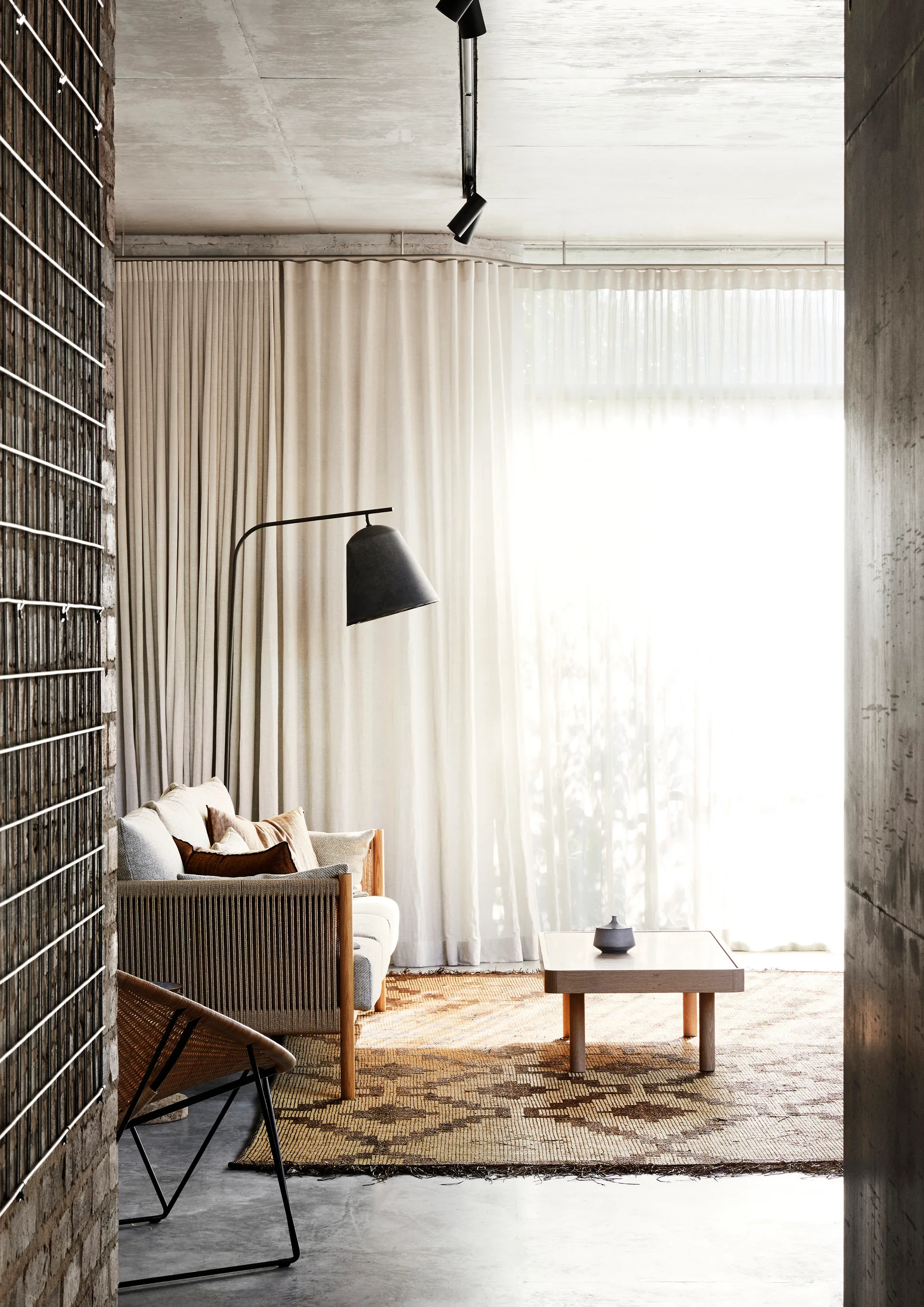 A bright living area features a woven-sided sofa, low wooden coffee table, and patterned rug set against floor-to-ceiling sheer drapes.
