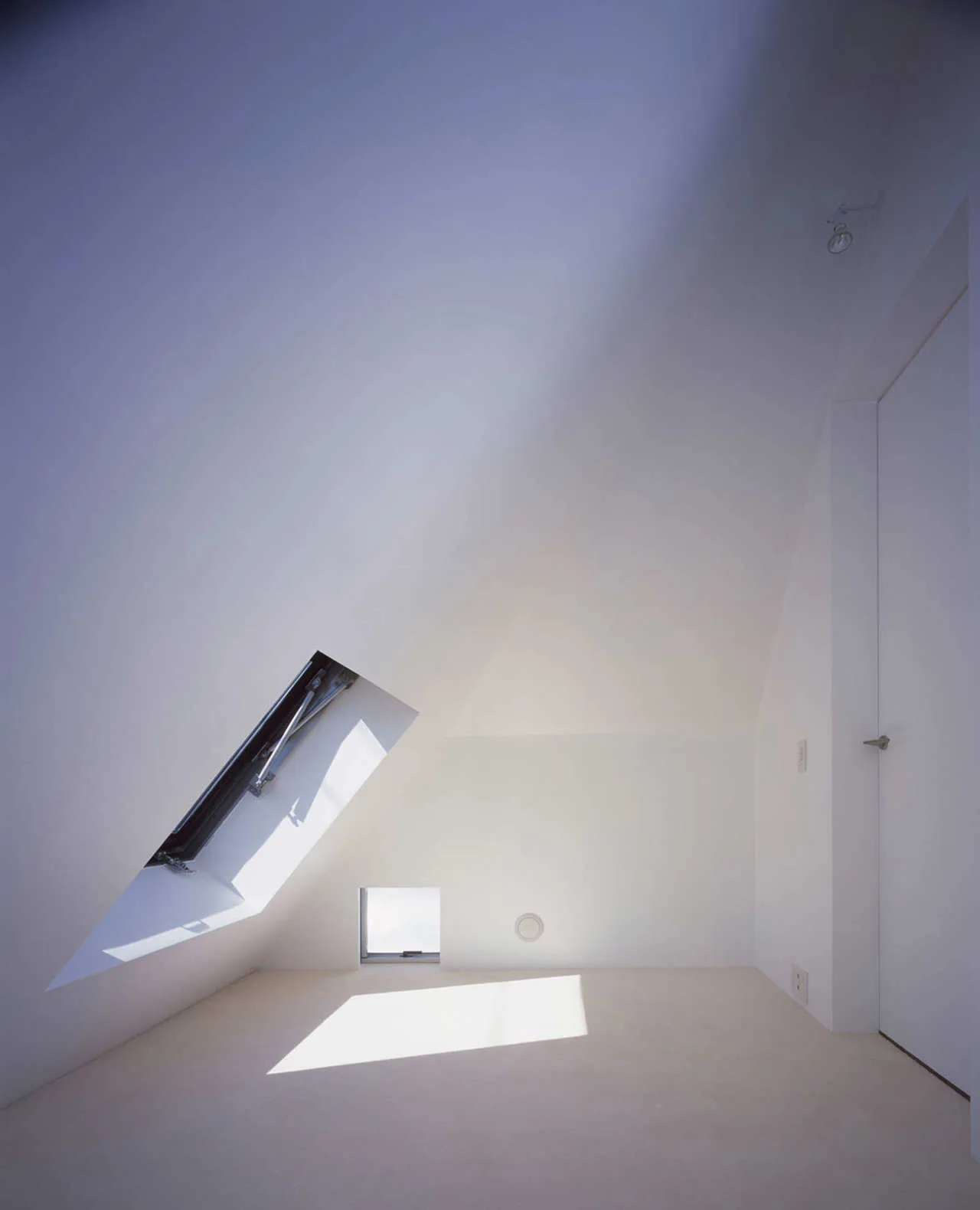 A minimalist empty attic room with white walls, a sloped ceiling, an open skylight, and a small square window near the floor.
