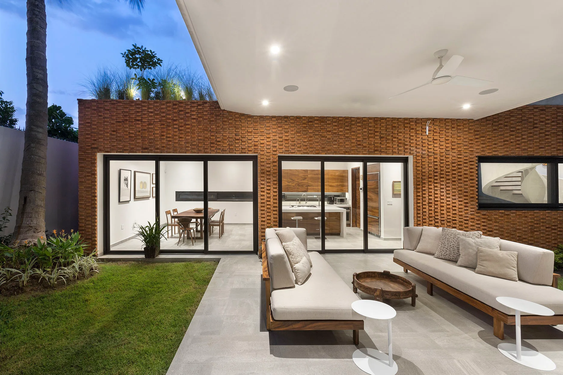 Two neutral outdoor sofas sit on a grey stone tile patio under a white ceiling overhang, set against a highly textured reddish-brown brick wall.
