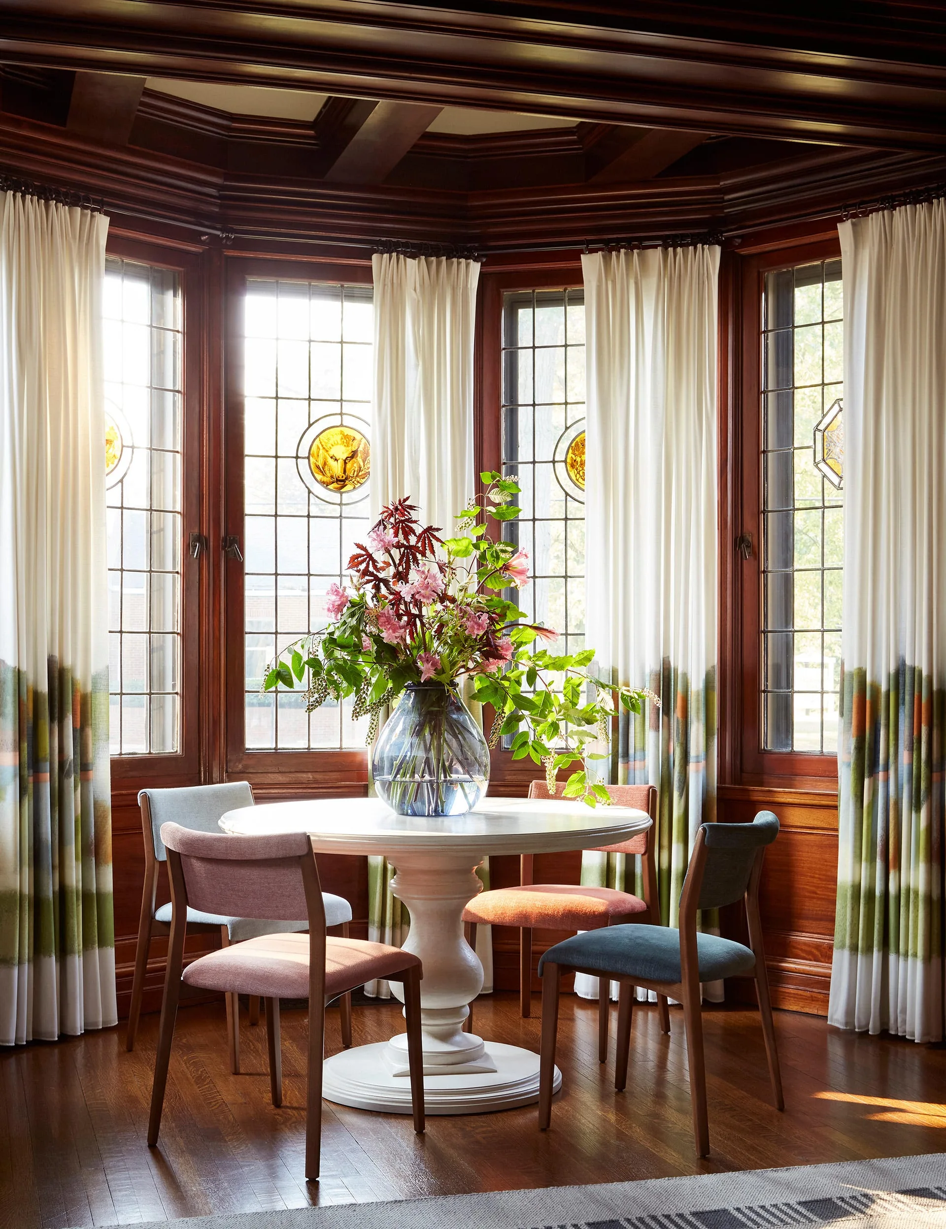 A round white pedestal table with multi-colored dining chairs is placed inside a dark wood-paneled bay window nook.