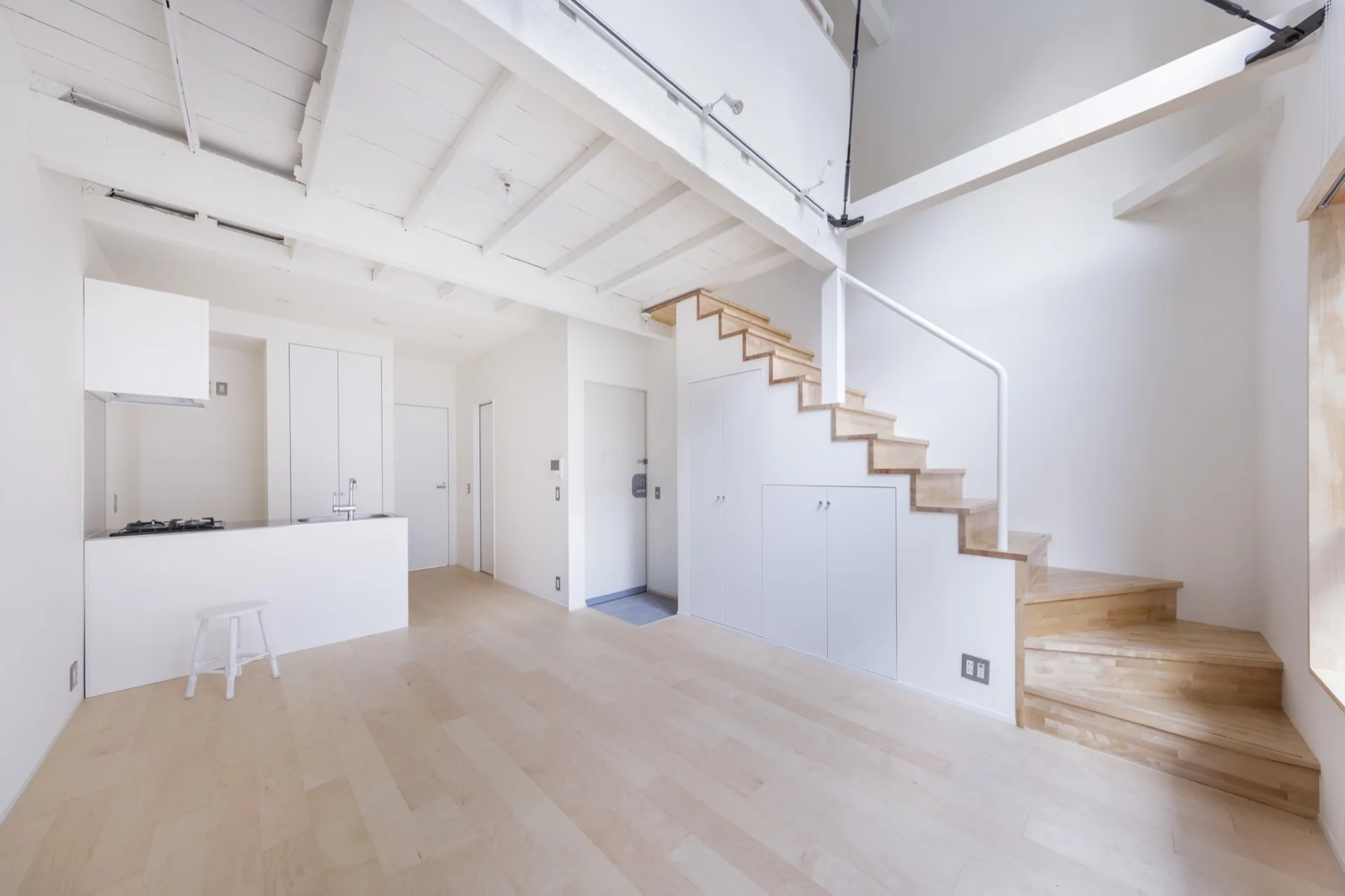 A minimalist white open-plan living area featuring light wood flooring, a kitchen counter, and a wooden staircase with integrated storage below.