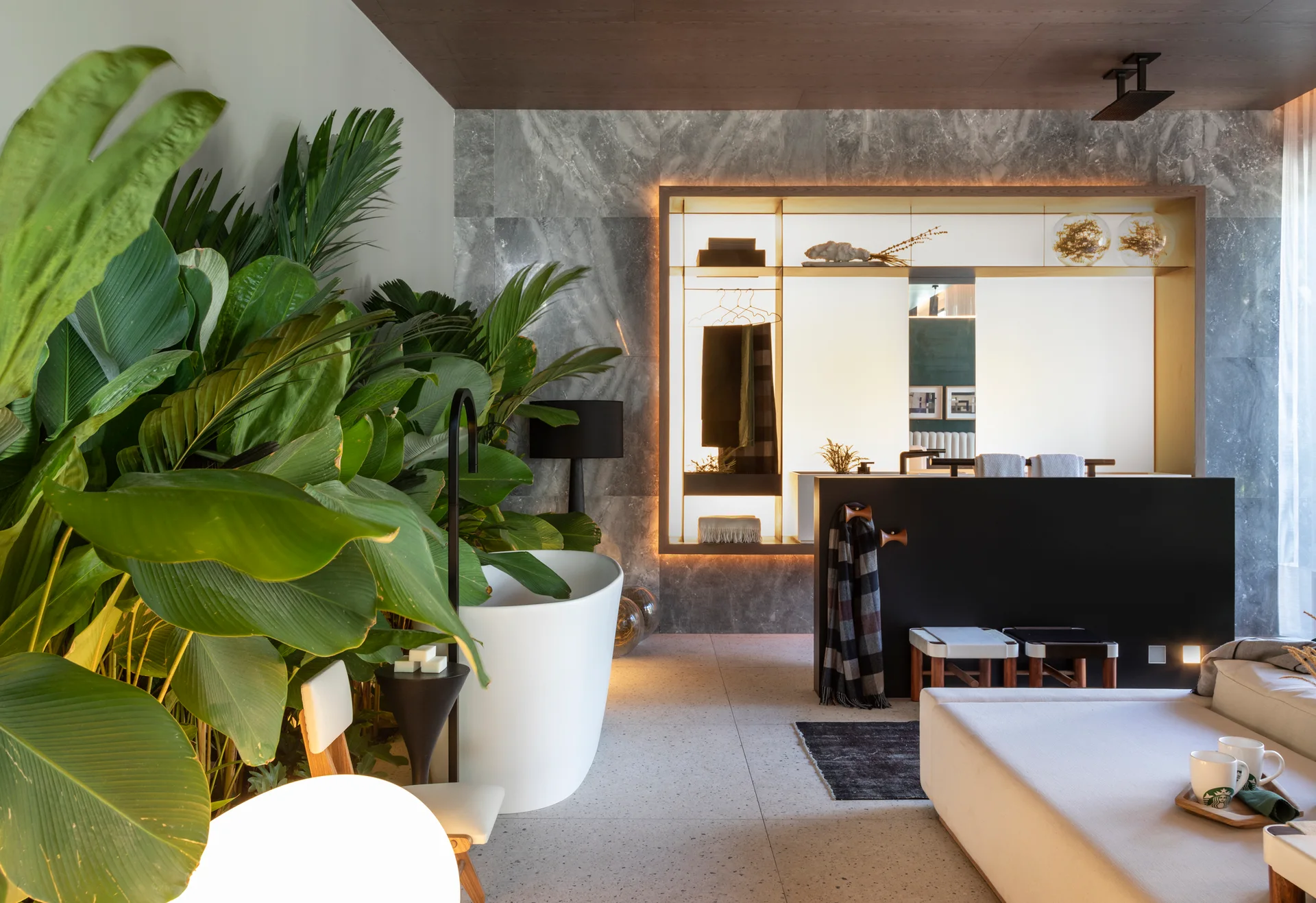 A modern bathroom features a white freestanding tub nestled in dense green plants, a black partition, and backlit shelving on a grey stone wall.