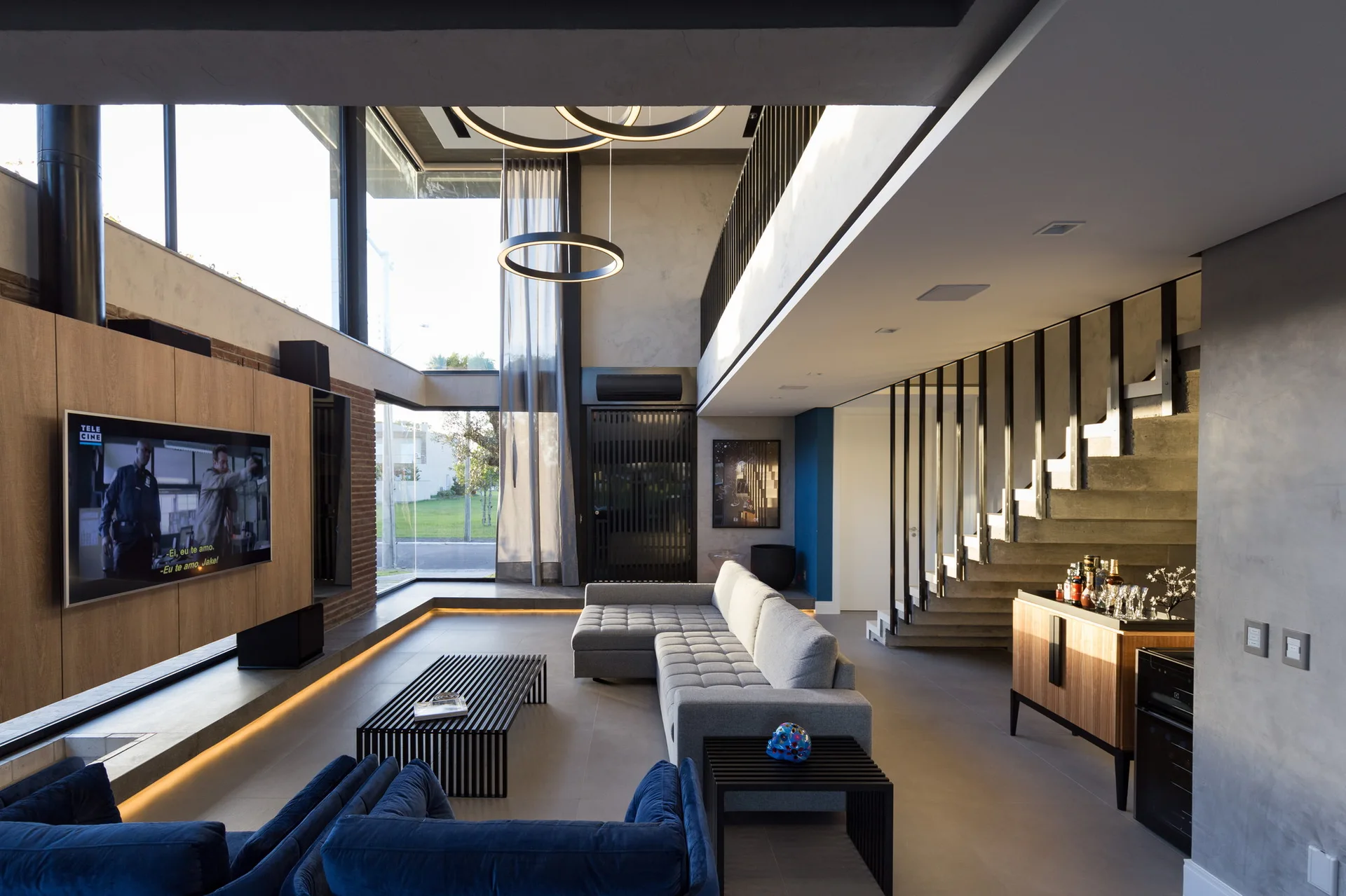 A spacious modern living room featuring a grey sectional sofa, slatted coffee table, concrete staircase, and large circular pendant lights.
