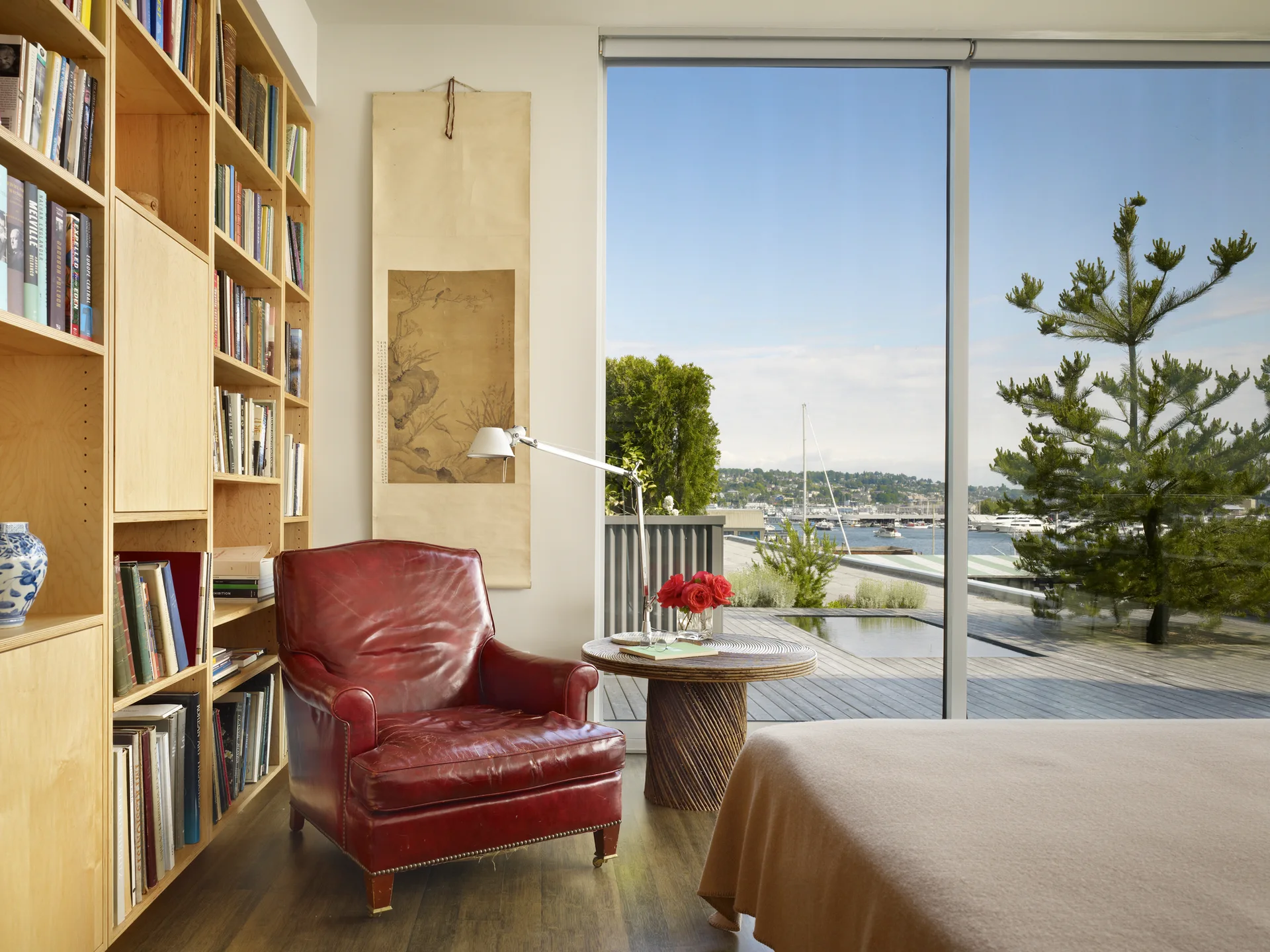 A red leather armchair sits beside a built-in light wood bookcase next to a large window overlooking a marina.