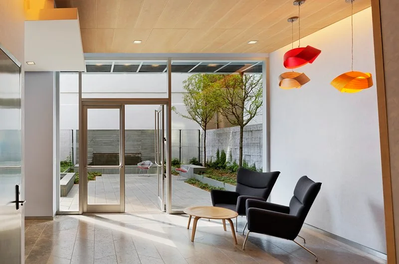 Two dark grey armchairs and a small round wooden coffee table sit in a modern lobby with a wood ceiling, facing a glass door that opens to a courtyard.